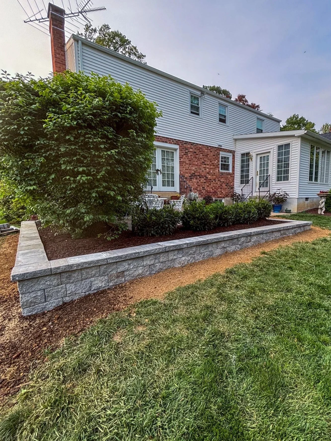 A backyard garden with a brick and gray stone retaining wall, green grass in the foreground, and a house with brick and siding exterior in the background.