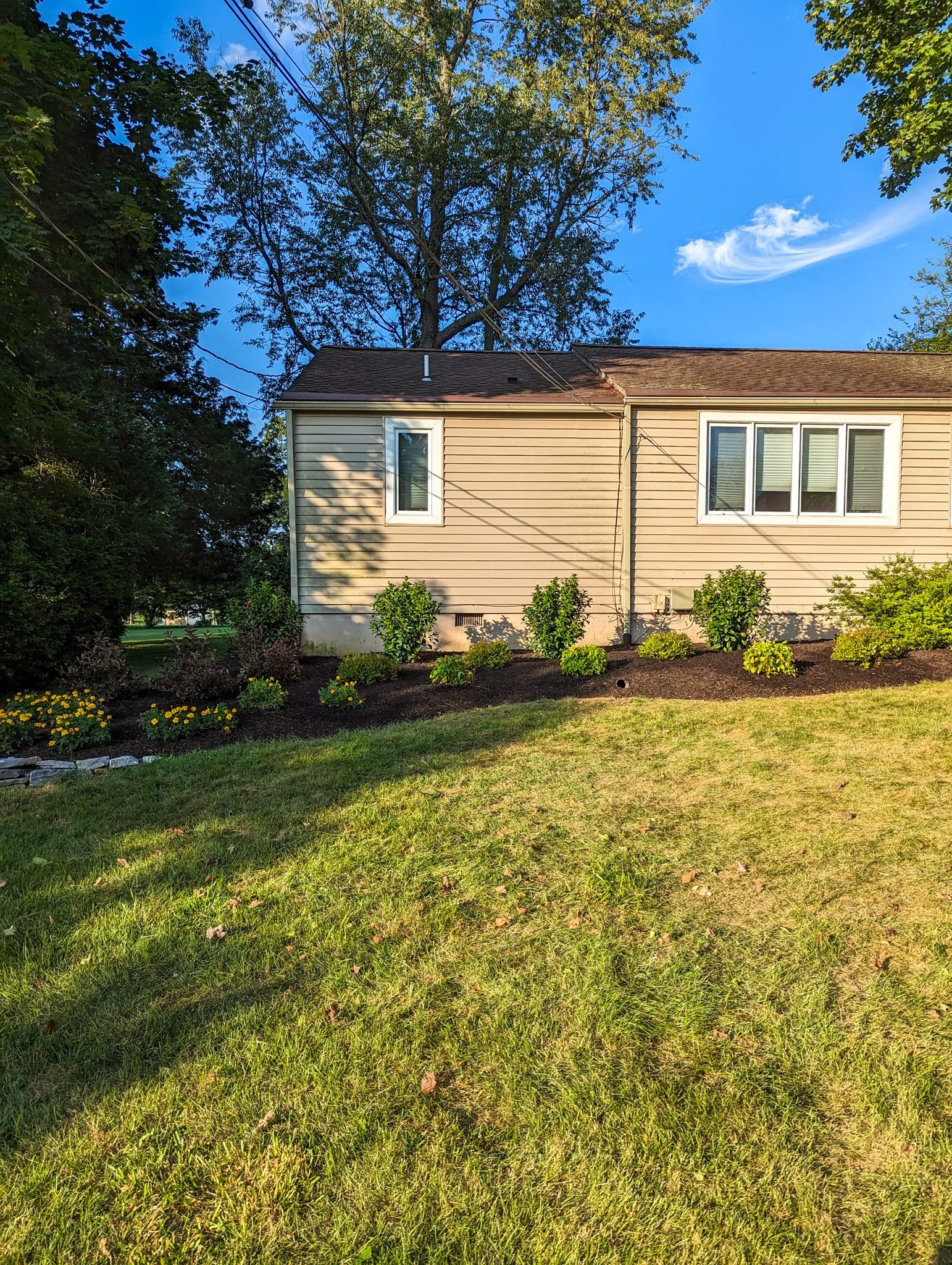A beige house with white window trims, surrounded by a well-maintained garden with bushes and flowers, under a blue sky with some clouds and a tall tree in the background.