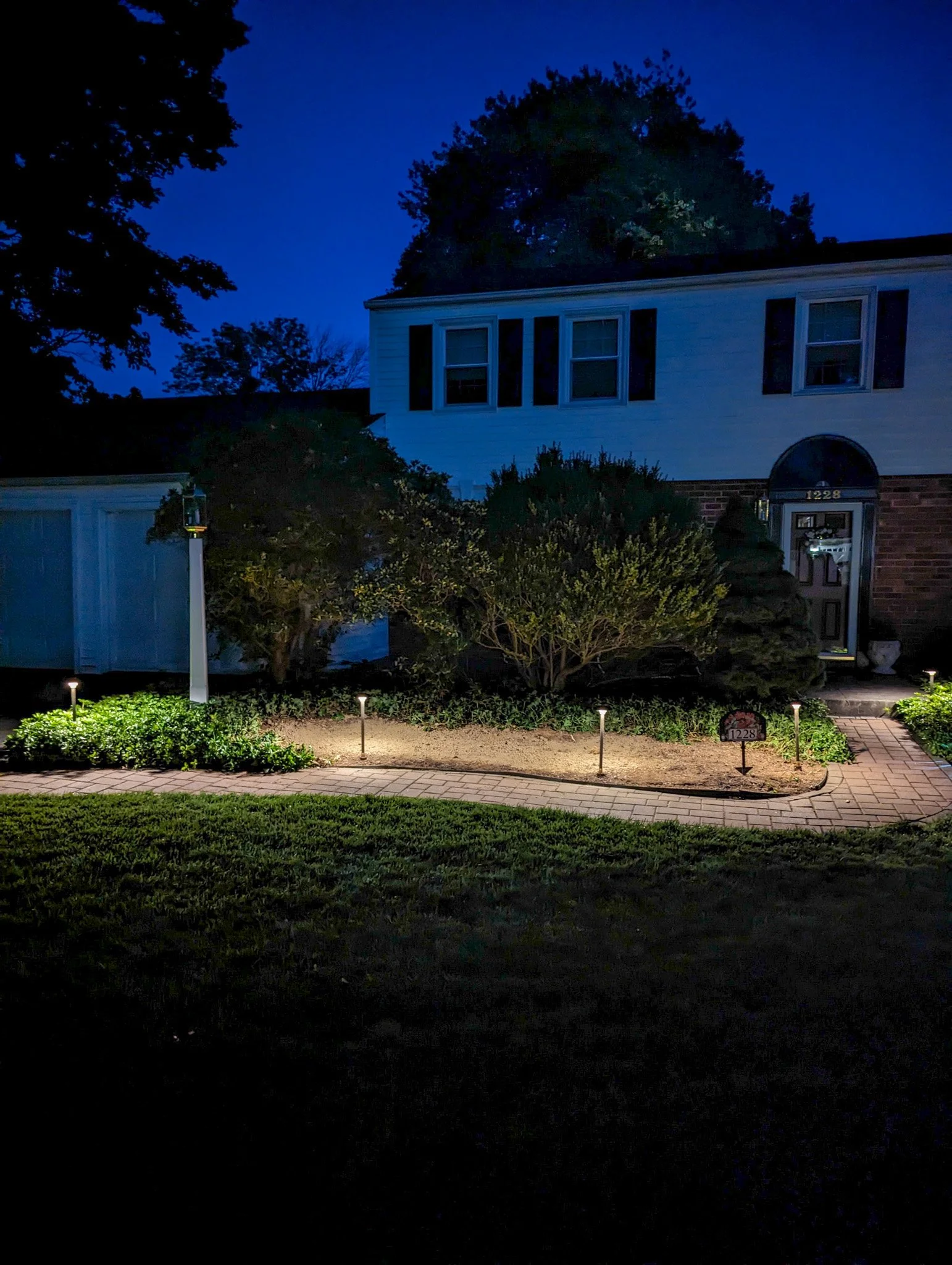 A residential house at night with landscape lighting illuminating the front yard pathway, bushes, and trees.