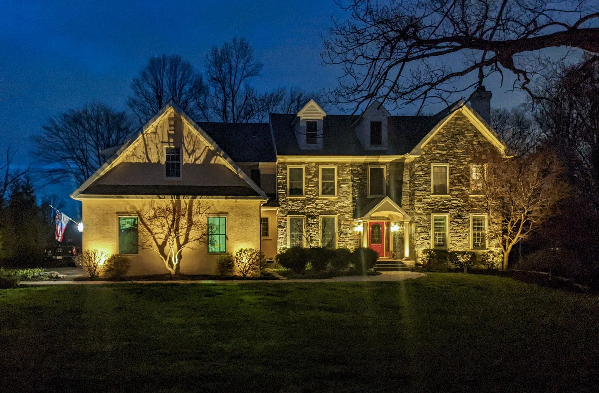 A large, illuminated house with stone and siding exterior, front porch, multiple windows, and a red door, surrounded by trees at night.