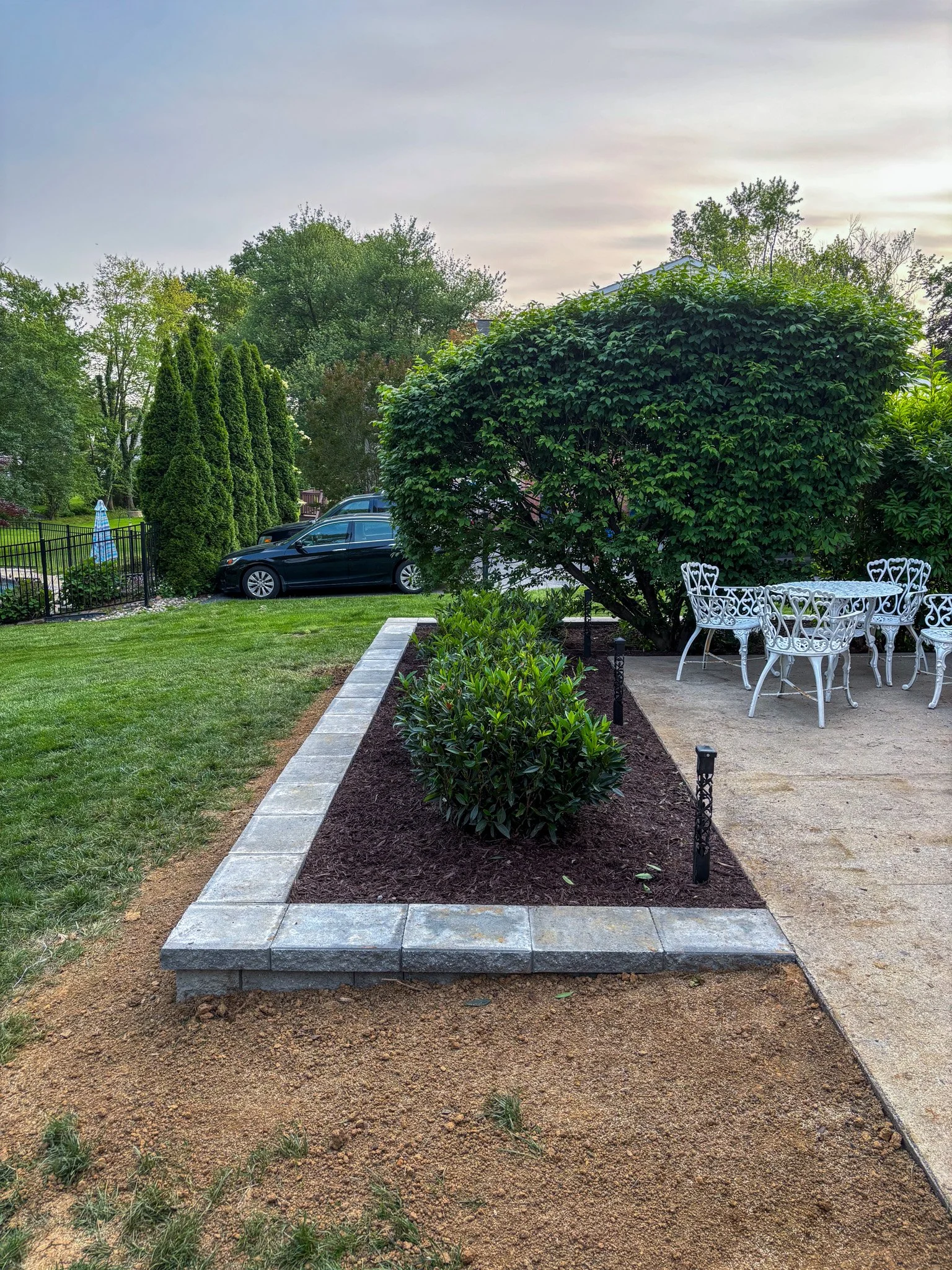 A landscaped backyard with a patio area containing white metal chairs and a table, a green bush, tiered window shrubs, a dining area, and a car parked in the background with trees and a cloudy sky.