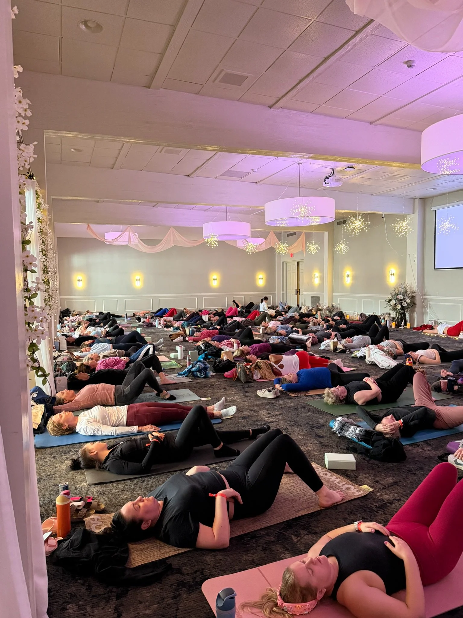 A large group of people practicing yoga or meditation in a spacious, softly lit indoor room with ceiling lights and draping decor.