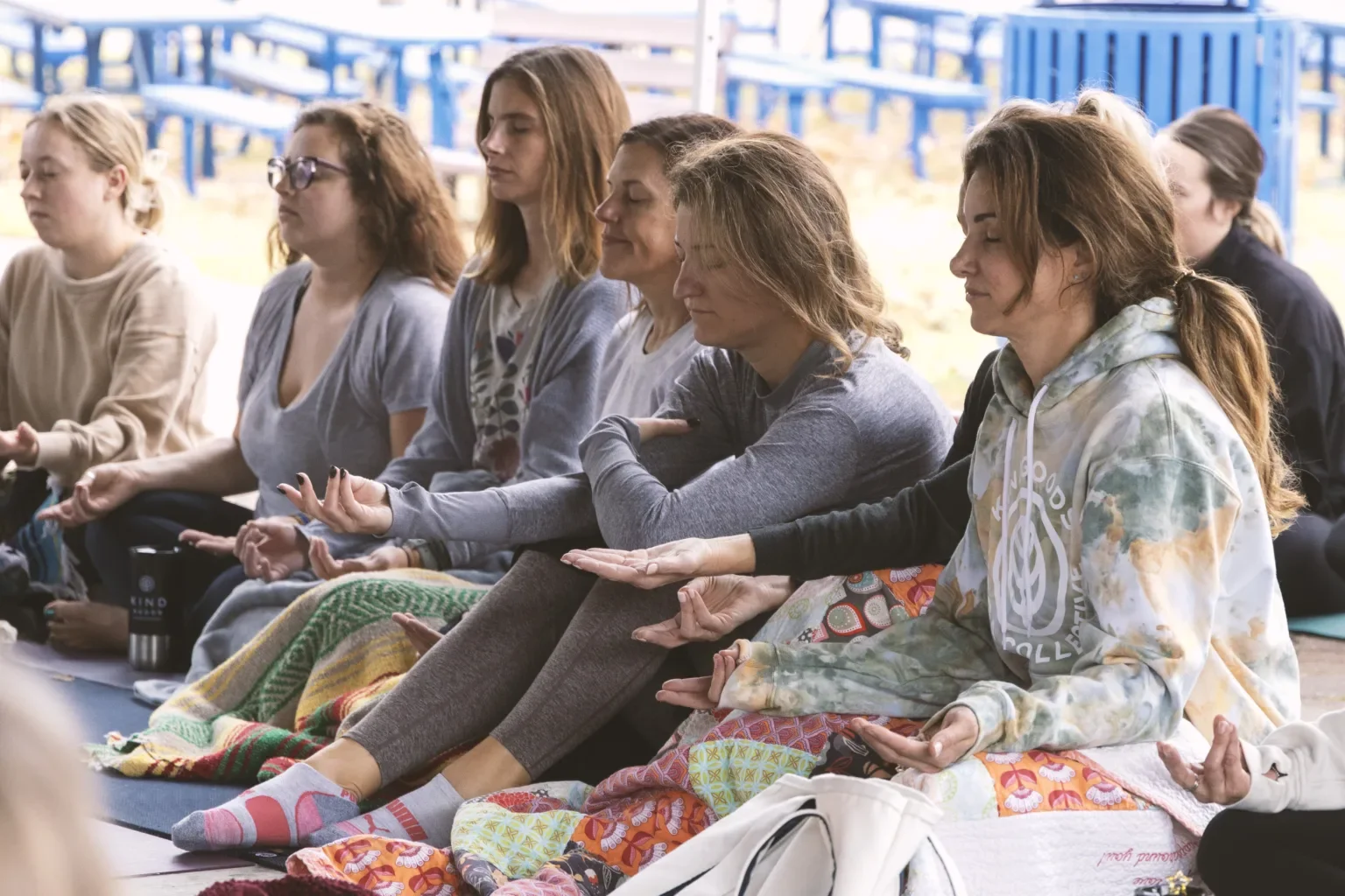 A group of women sitting cross-legged with eyes closed, meditating outdoors on a covered porch with blue chairs in the background.