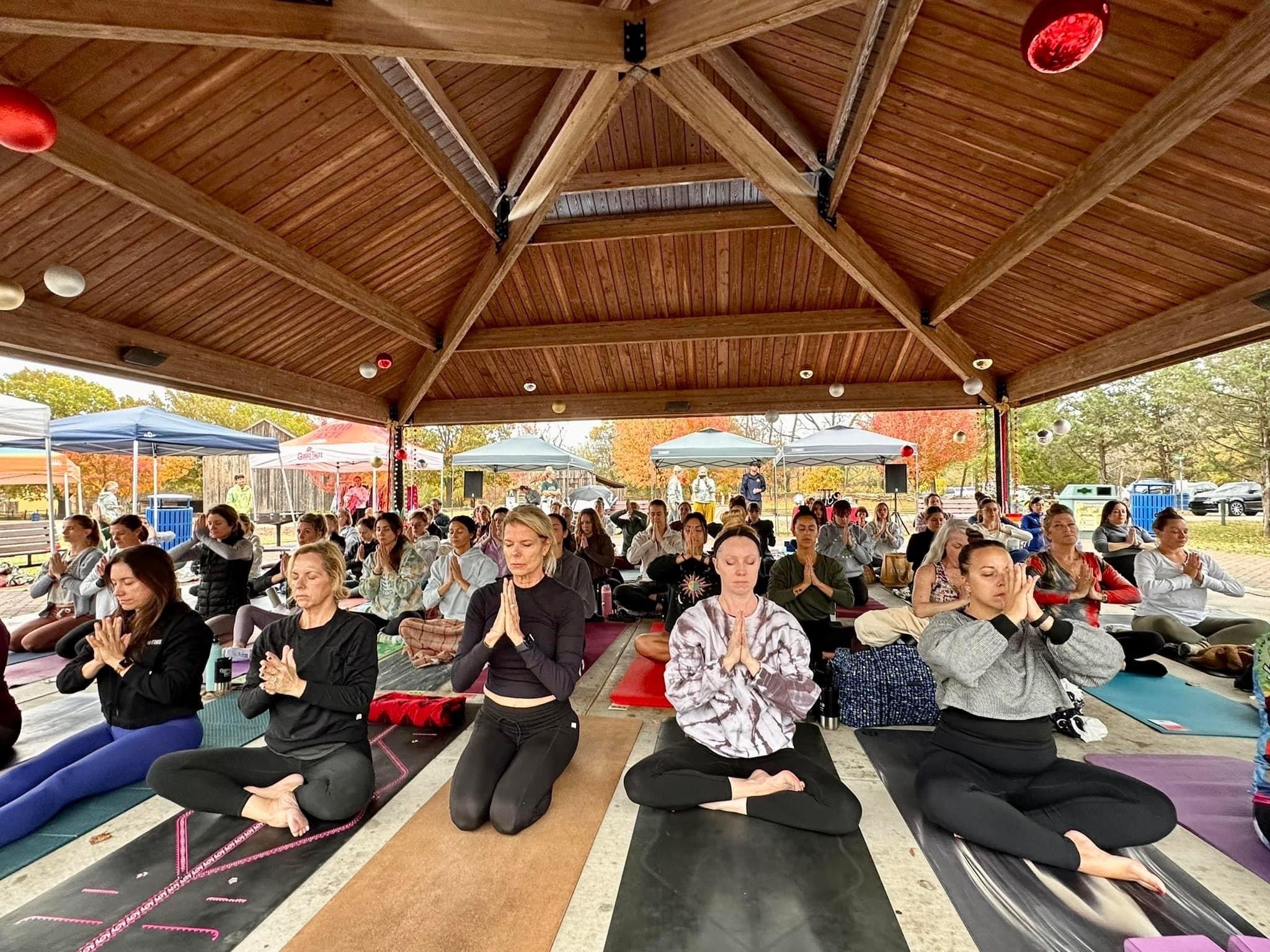 Group of people participating in outdoor yoga or meditation session under a wooden pavilion, seated on mats with hands in prayer position, with trees and tents visible in the background.