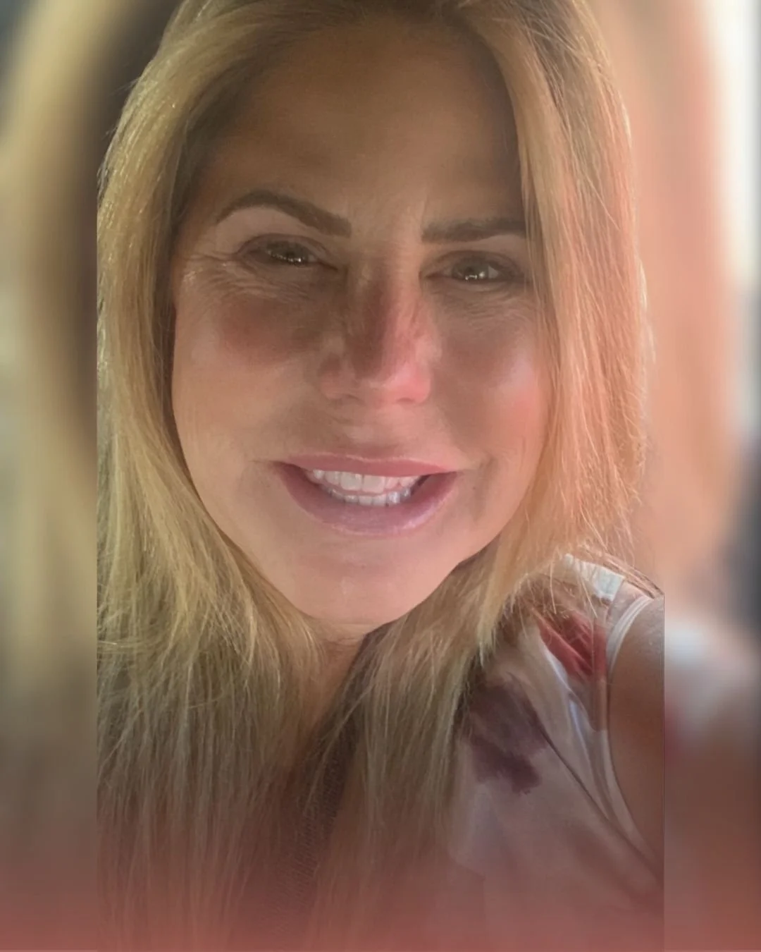 A woman with long blonde hair, smiling at the camera with visible teeth and some wrinkles around her eyes.