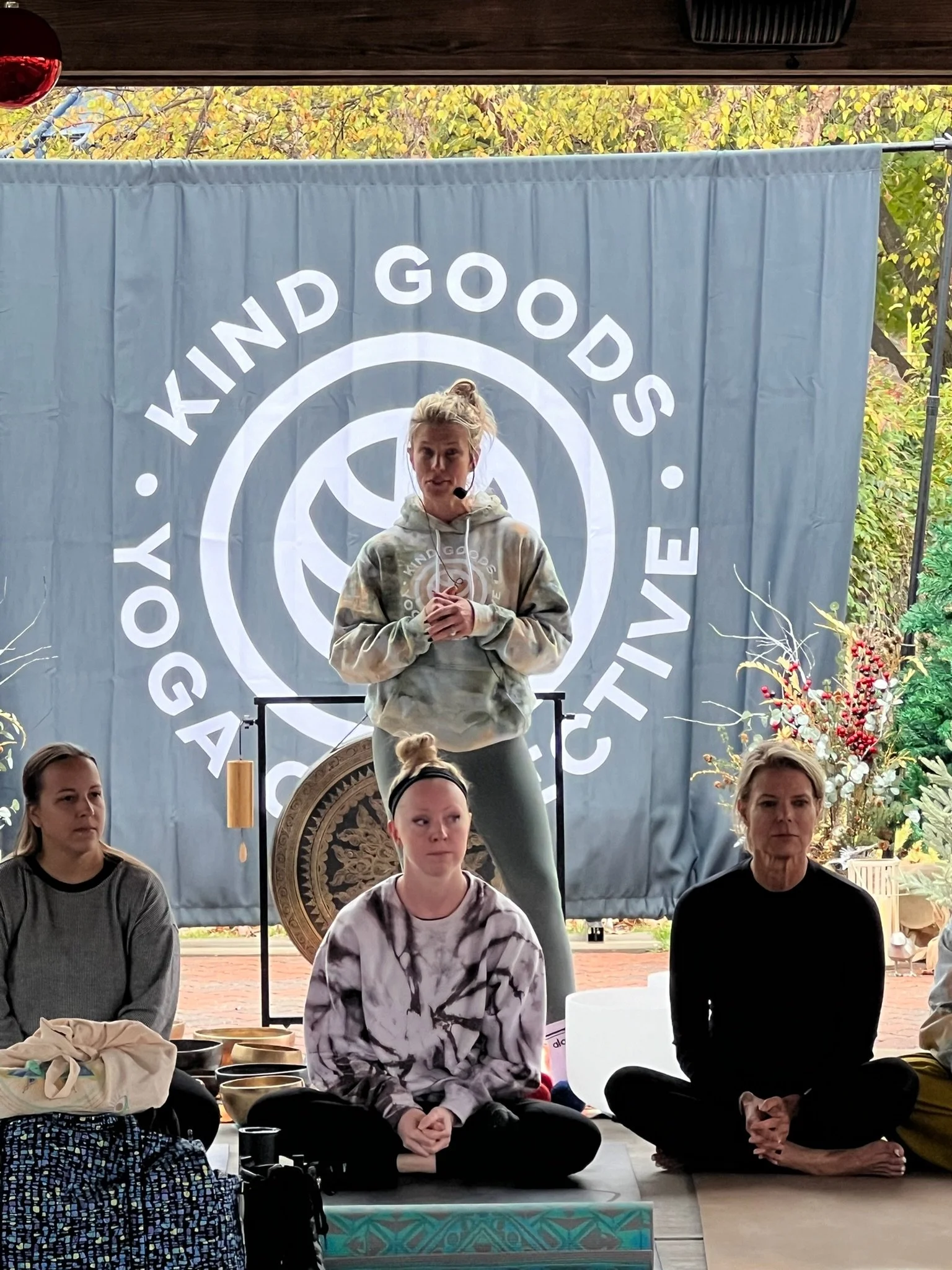 A woman standing on a stage or platform, wearing a hoodie and speaking into a microphone, in front of a large banner with the words 'KIND GOOD'S YOGA ACTIVE' and a logo. Several women are seated on the floor in yoga or meditation poses, with various items and decorative plant arrangements nearby.