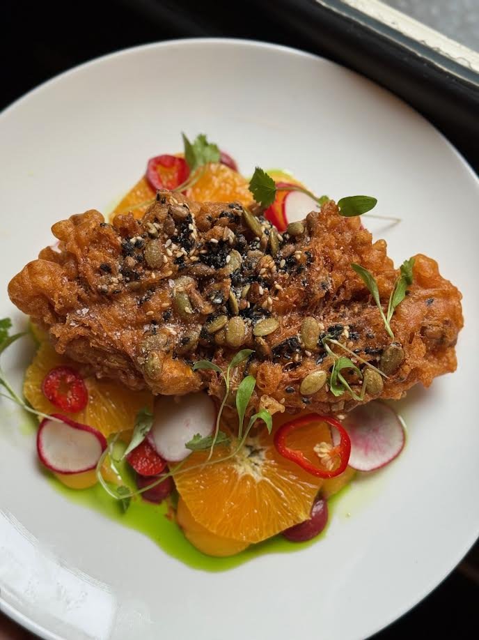Fried chicken topped with pumpkin seeds, served with orange slices, radishes, cherry tomatoes, microgreens, and colorful sauce on a white plate.