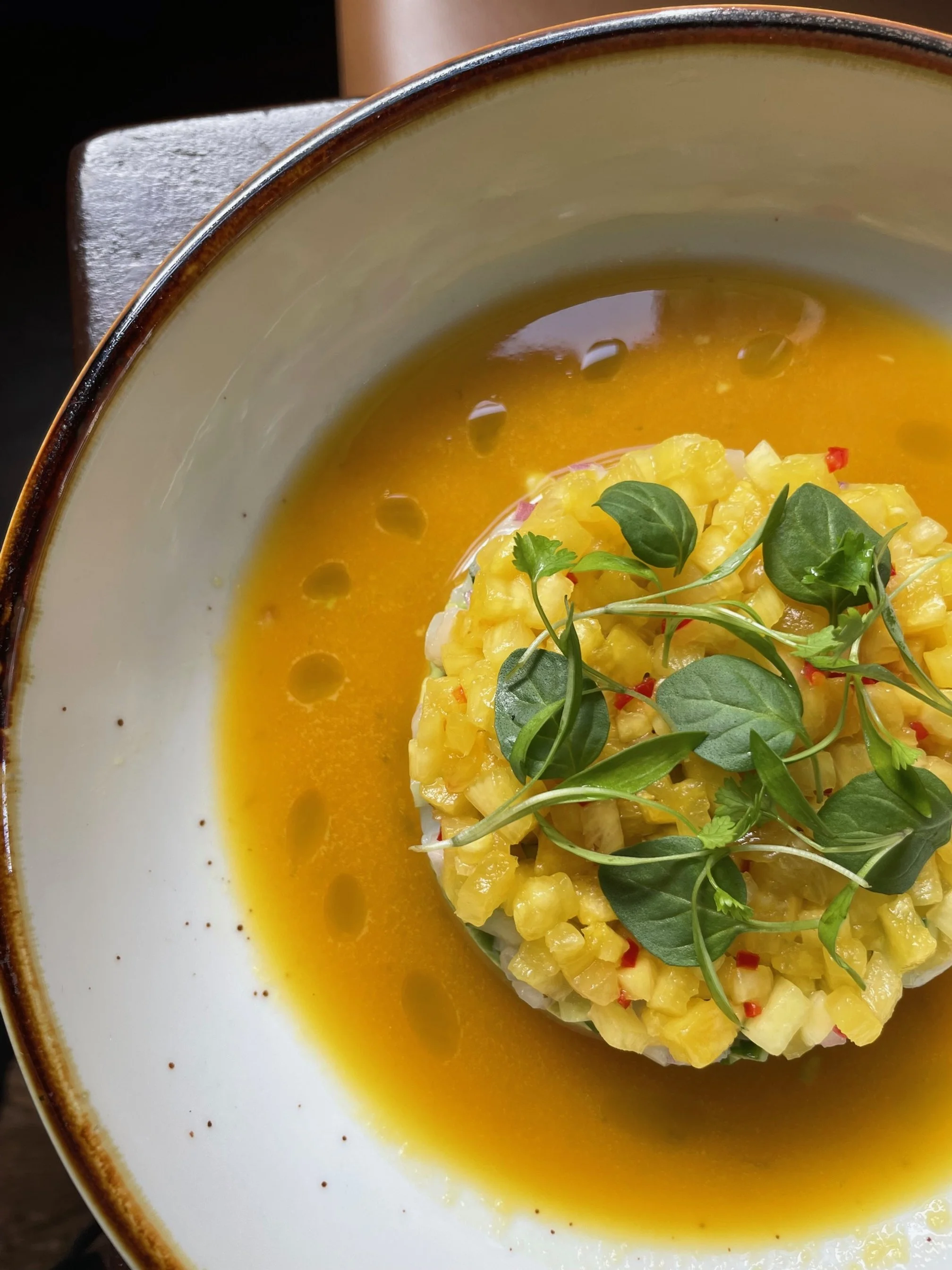 A bowl of yellow soup garnished with microgreens and a mound of diced fruit or vegetables at the center.
