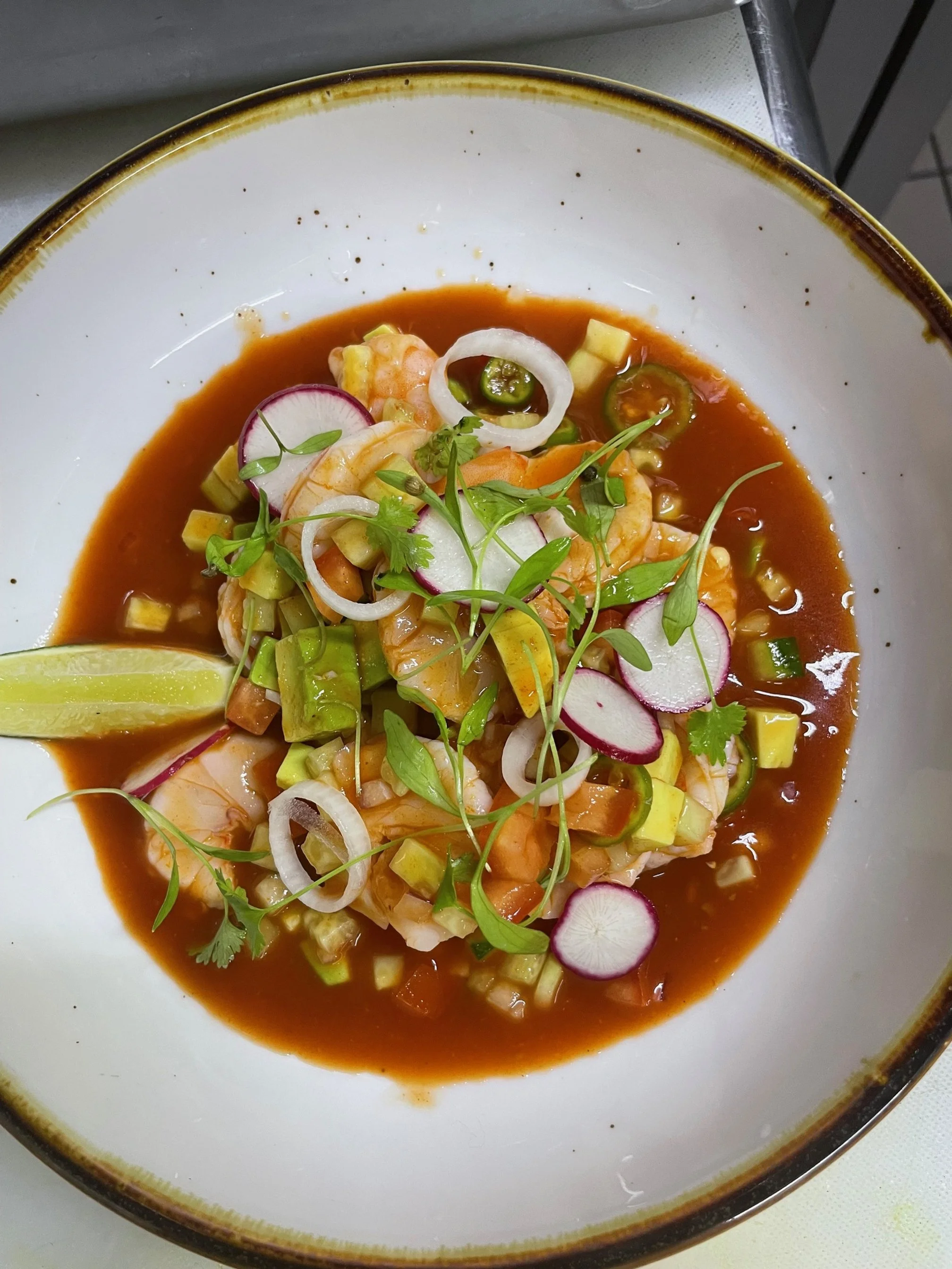 A bowl of shrimp ceviche garnished with radish slices, onion rings, chopped cucumber, cilantro, and a wedge of lime in a sauce.