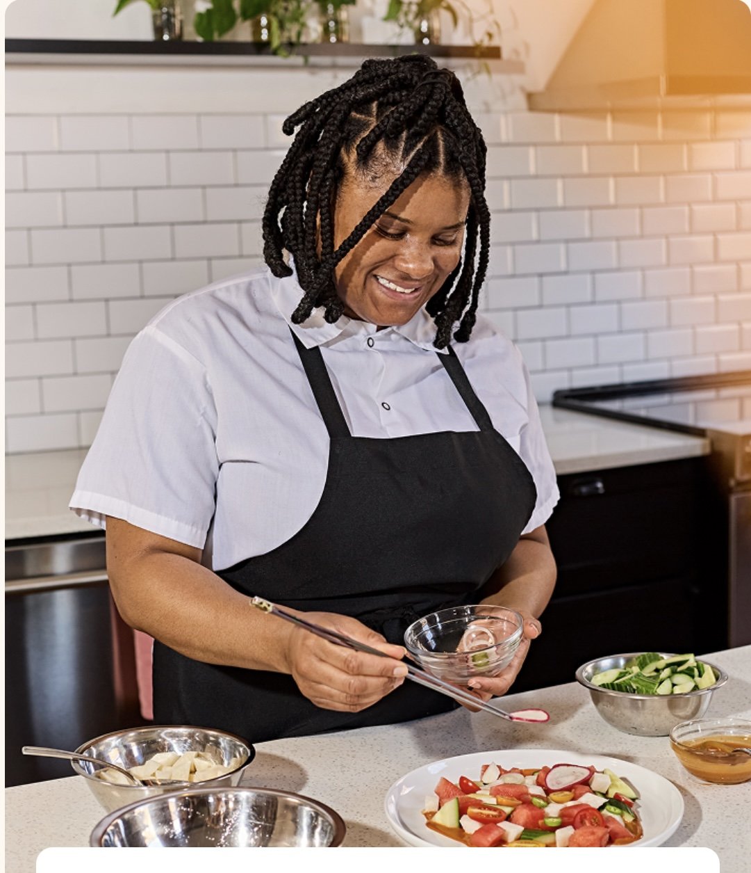 A woman with braided hair smiling while preparing a salad in a modern kitchen. She is wearing a white shirt and a black apron, holding a small bowl and a spoon. On the counter are bowls with vegetables and dressing.