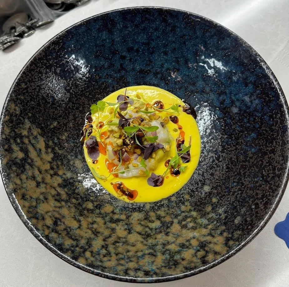 A gourmet dish featuring a yellow sauce, garnished with microgreens, crushed nuts, and balsamic glaze, served in a black patterned bowl.