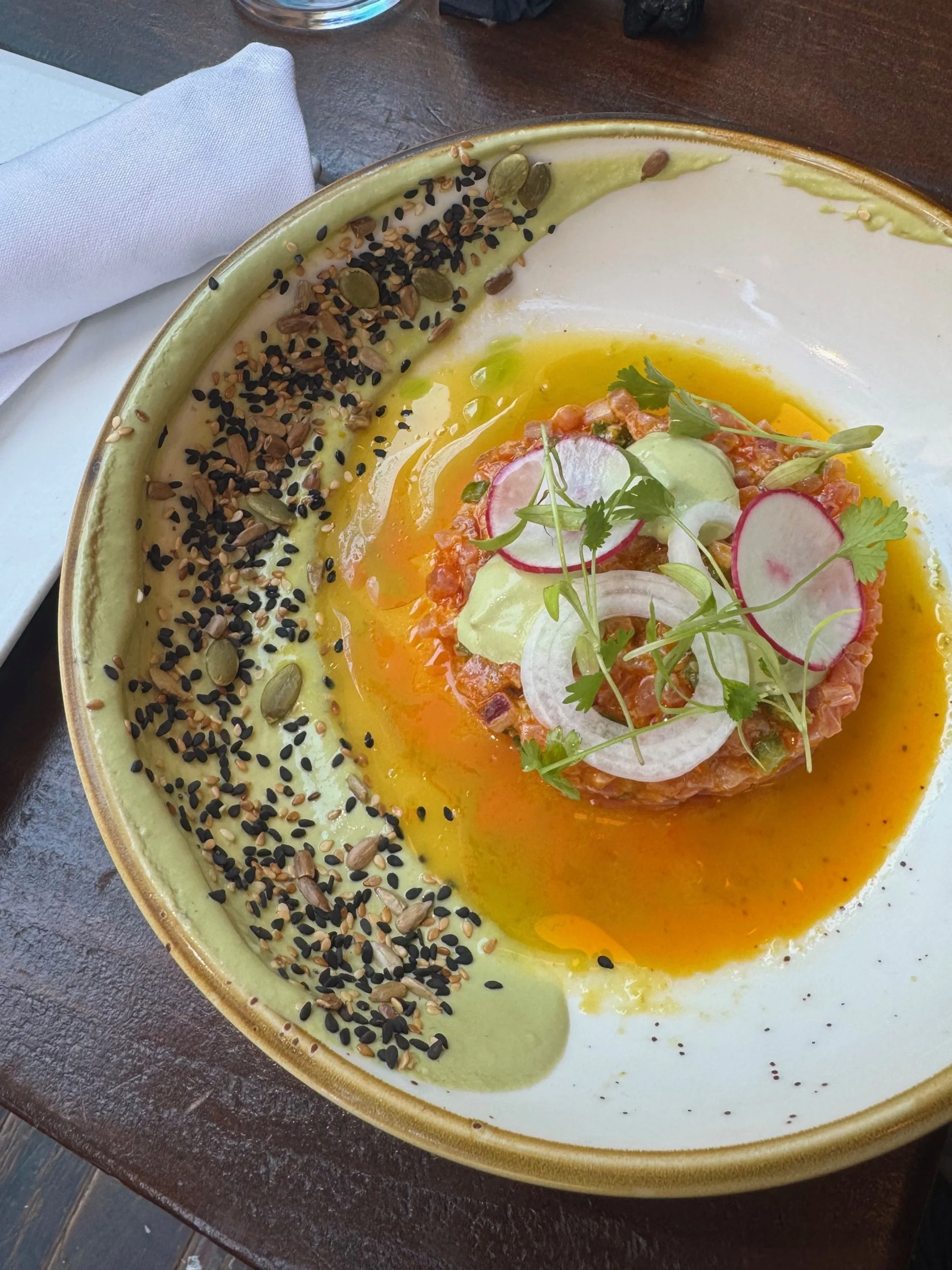 A plate of tuna tartare garnished with radish slices, onion, herbs, and microgreens, surrounded by a green sauce and sprinkled with mixed seeds on a table.