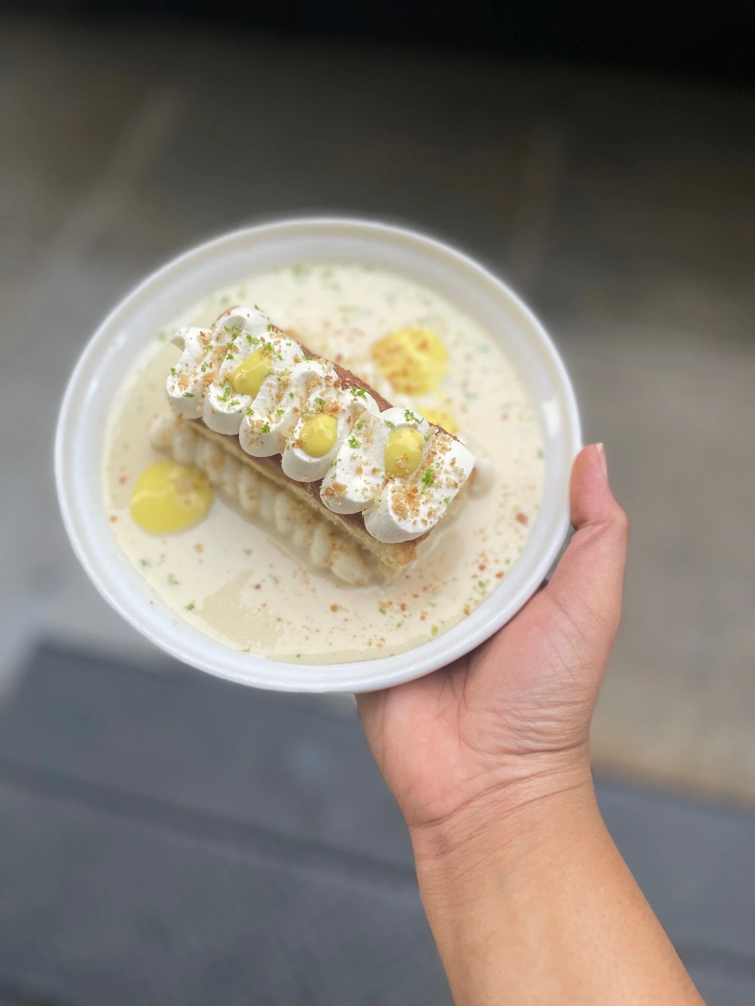 A hand holding a bowl with a layered dessert topped with whipped cream, lime zest, and yellow garnish, sitting in a creamy sauce.