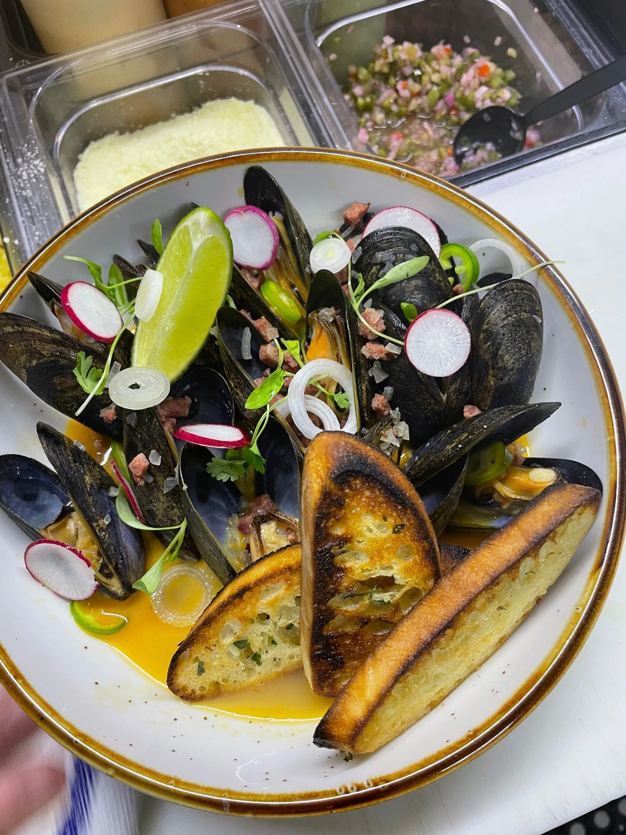 A bowl of cooked mussels garnished with sliced radishes, green onions, cilantro, and a lime wedge, with toasted bread slices and a side of chopped relish in the background.