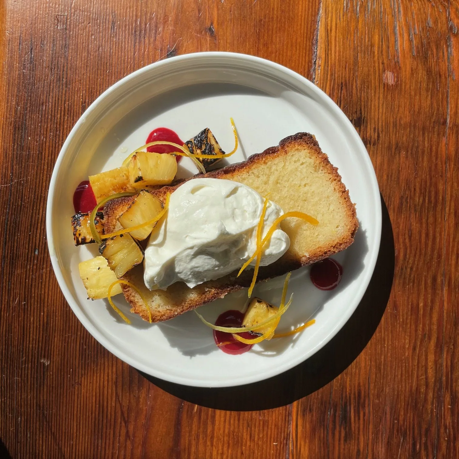 A plate of French toast topped with whipped cream, grilled pineapple, and garnished with lemon zest and cherry sauce, served on a white round plate on a wooden table.