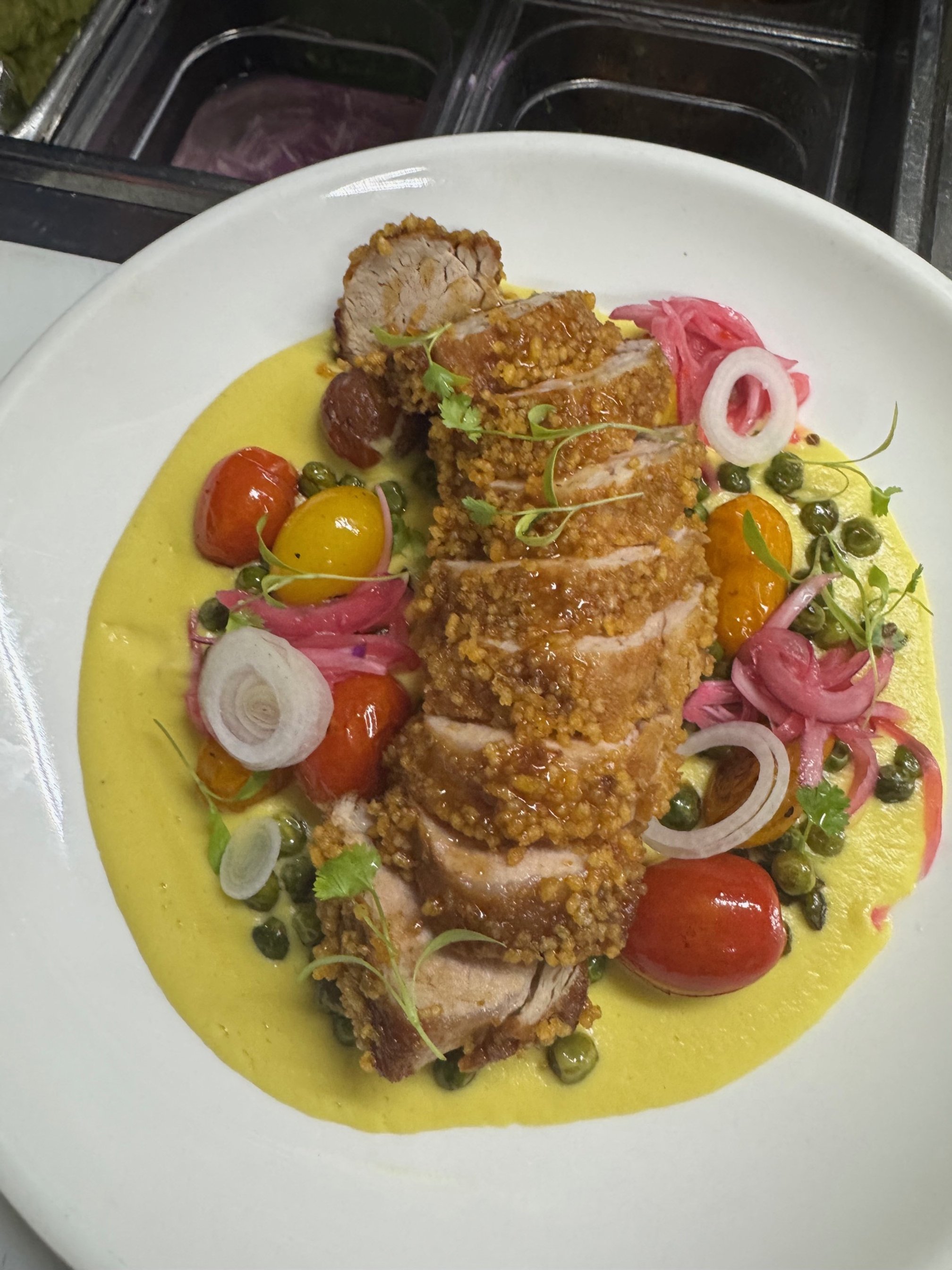 Fried chicken breast slices served with yellow sauce, cherry tomatoes, pickled onions, pink pickled vegetables, green peas, and garnished with cilantro on a white plate.