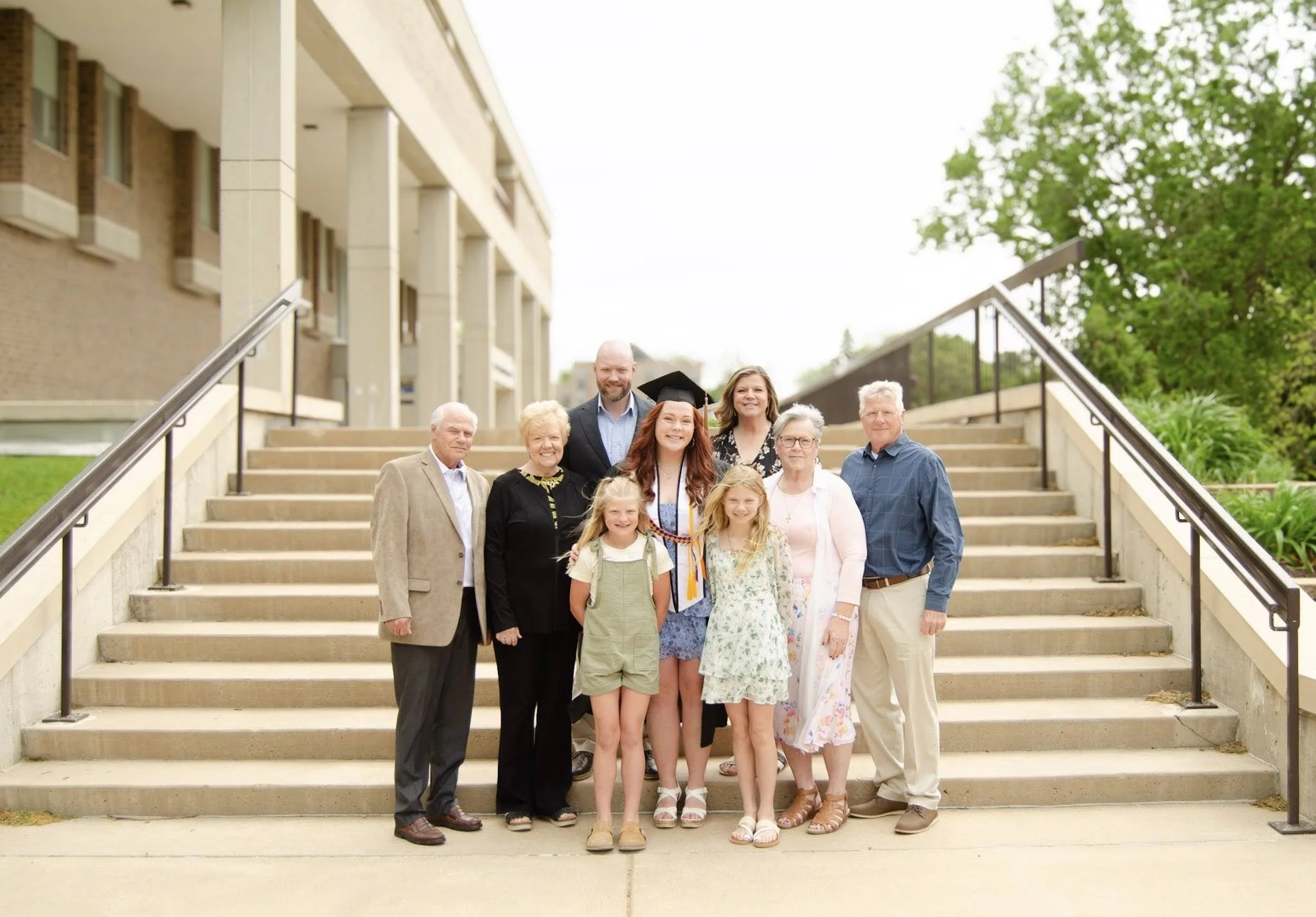 A graduation celebration with a young woman in a cap and gown surrounded by family members on outdoor stairs.