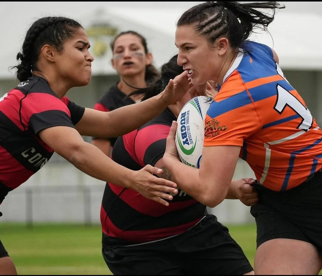 Two female rugby players, one in a black and red uniform and the other in an orange and blue uniform, are engaged in a tackle during a game. The woman in orange holds a rugby ball with the logo 'Big Rugby' while the woman in black and red tries to block her. A third woman in the background appears to be a referee or official.
