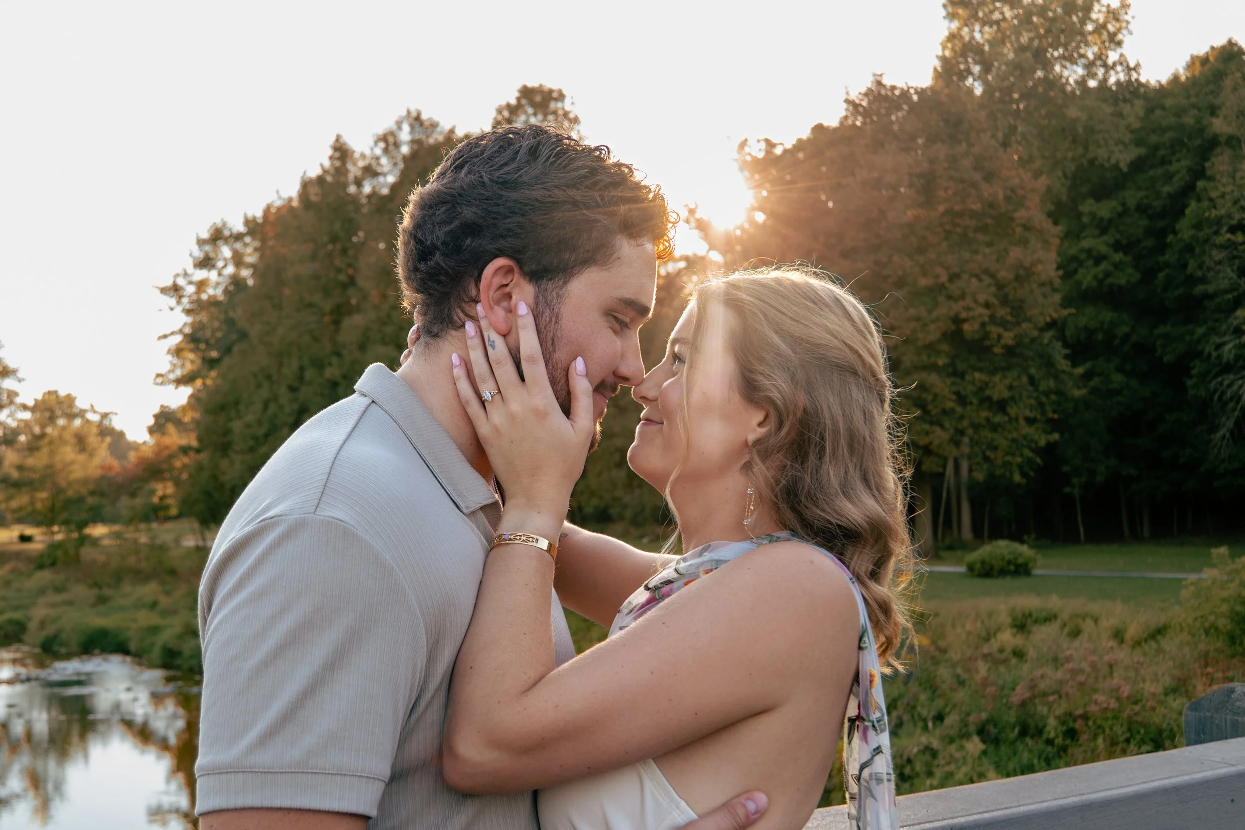 A couple embracing outdoors at sunset, with trees and a pond in the background.