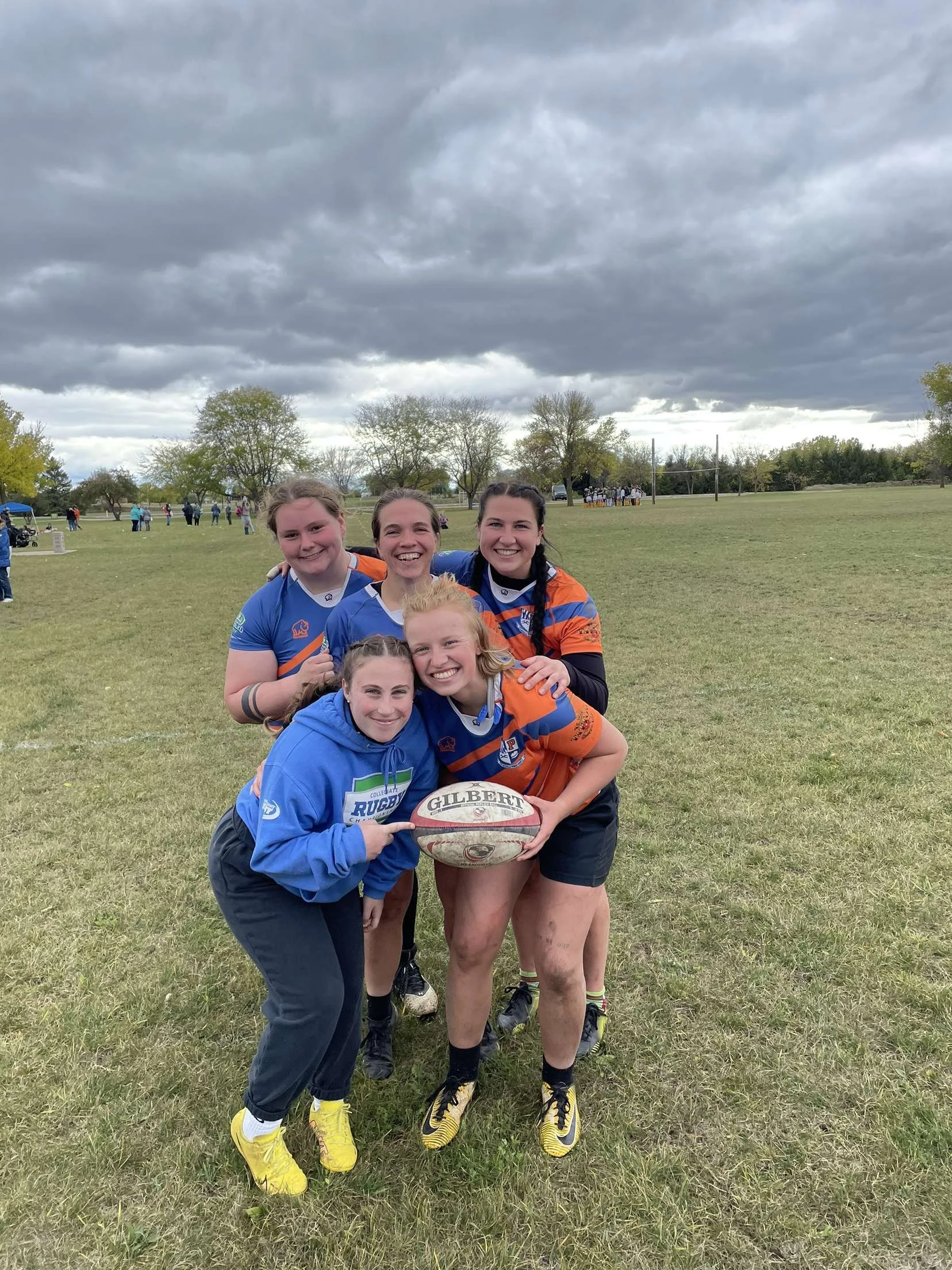 Five young women in rugby uniforms and casual clothes pose on a grassy field, smiling with one holding a rugby ball. Overcast sky, trees, and people in the background.