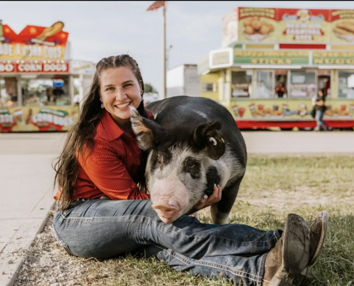A young woman with long dark hair, wearing a red shirt and jeans, sitting on the grass next to a large pig, with a carnival in the background.
