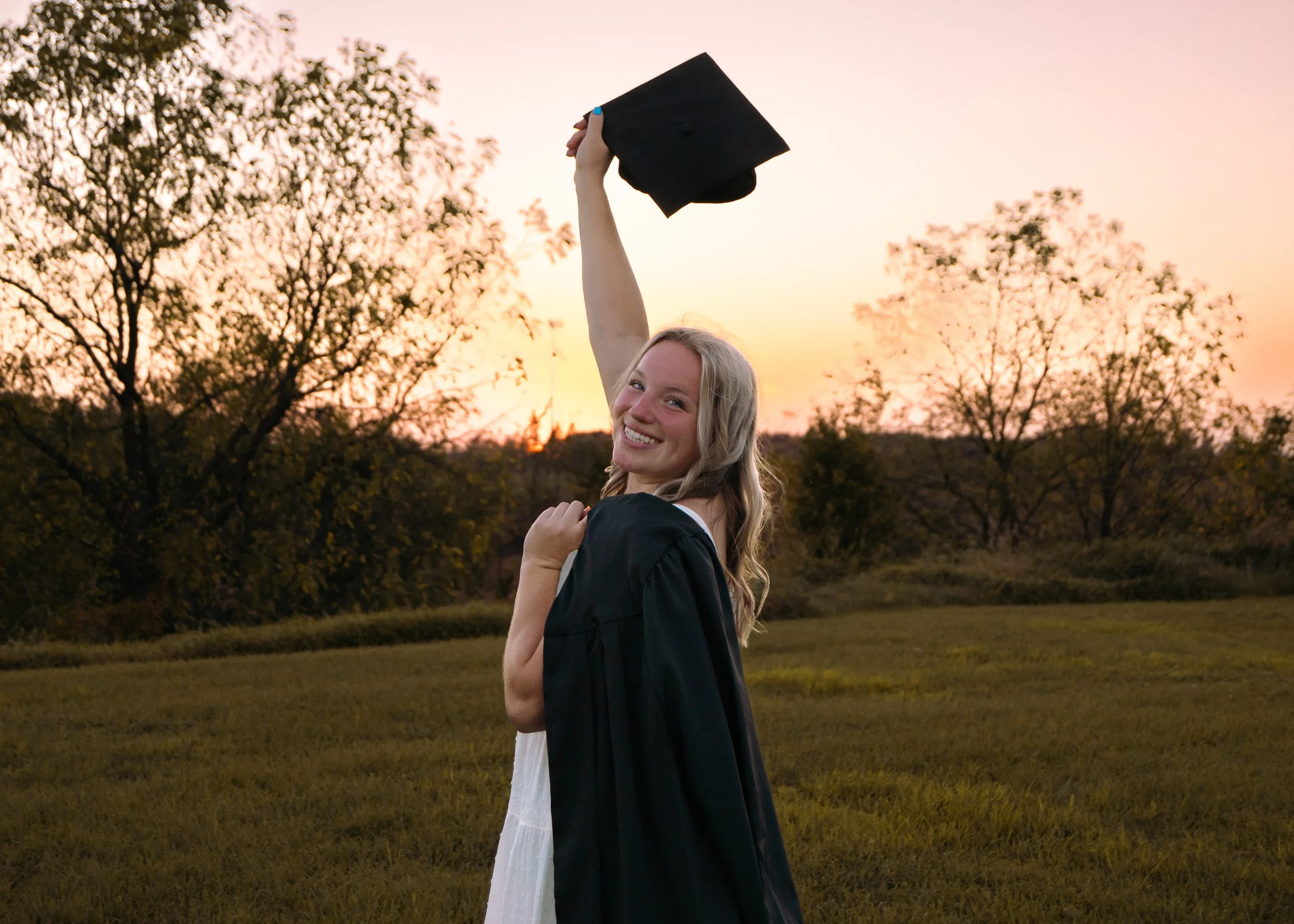 A young woman in a graduation gown smiling and holding her cap in the air outdoors during sunset.