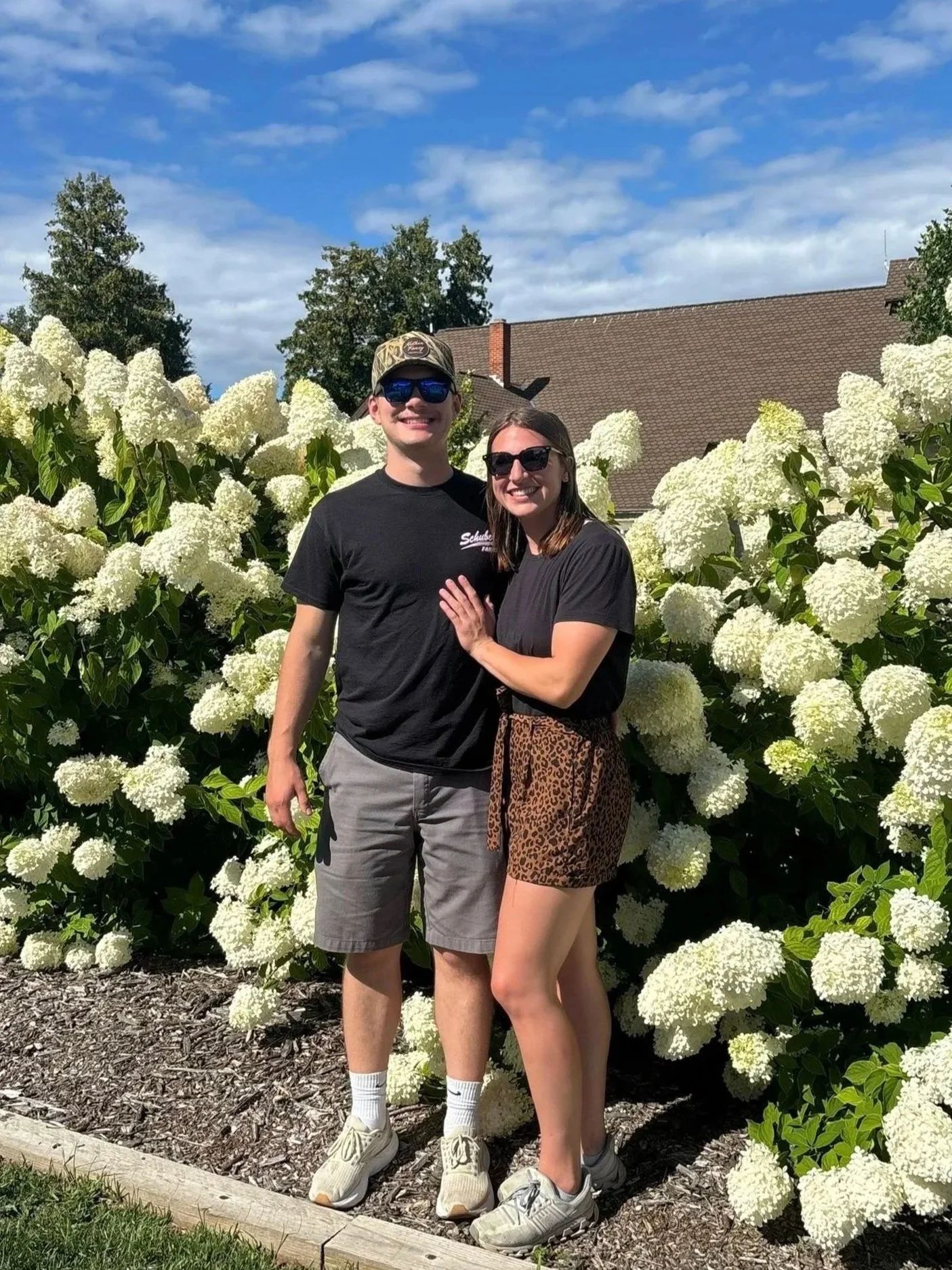 A young man and woman standing together outdoors in front of large white hydrangea flowers, smiling and wearing sunglasses on a sunny day.
