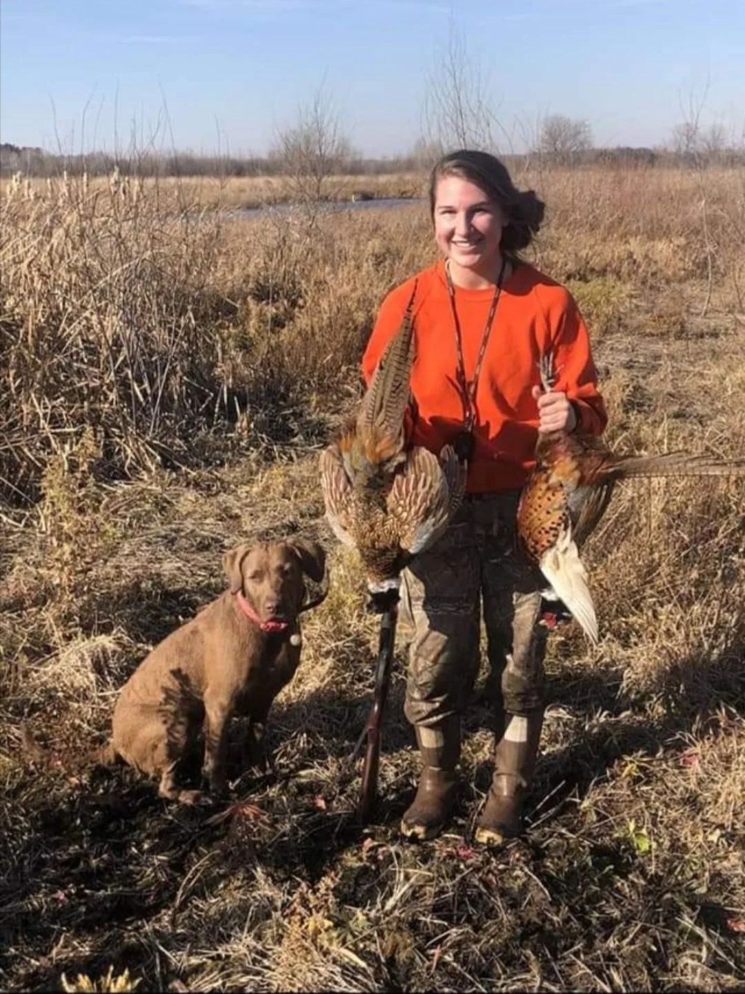 A woman smiling outdoors in a field holding two ducks, with a dog sitting beside her.