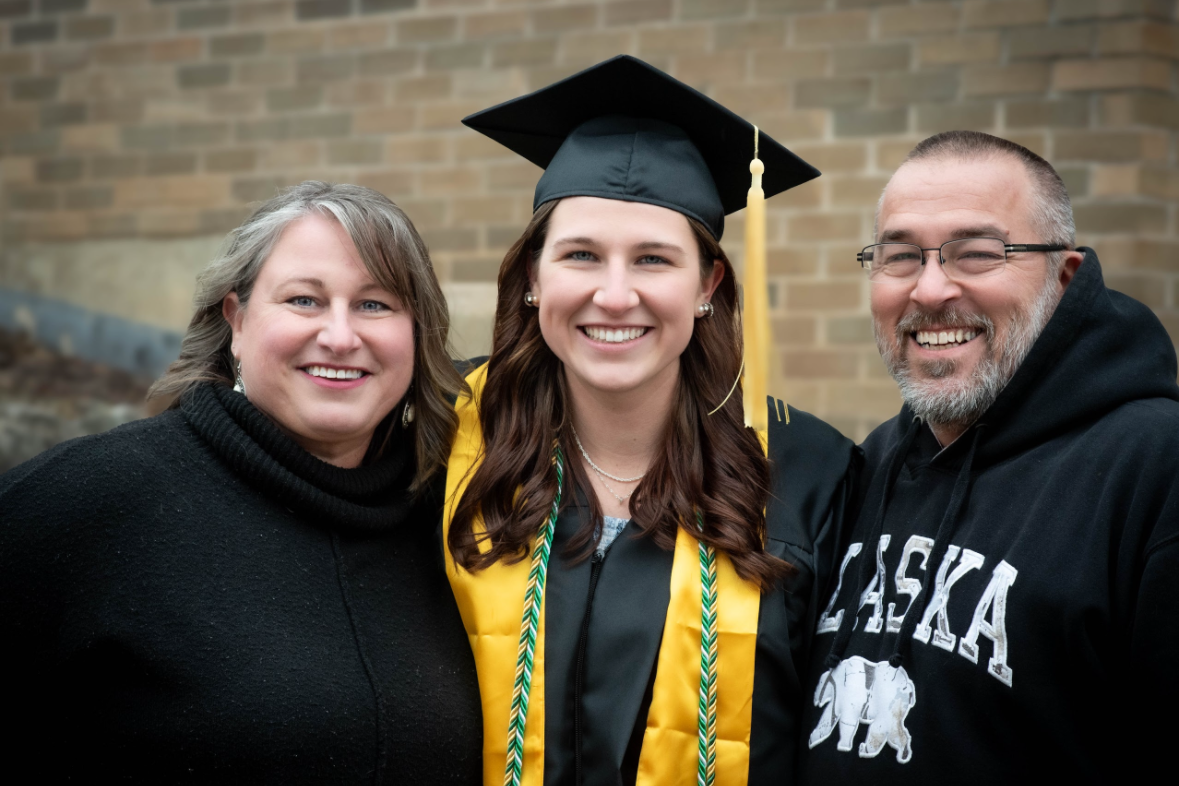 A young woman in a graduation cap and gown smiling with her parents who are on either side of her, standing in front of a brick wall.