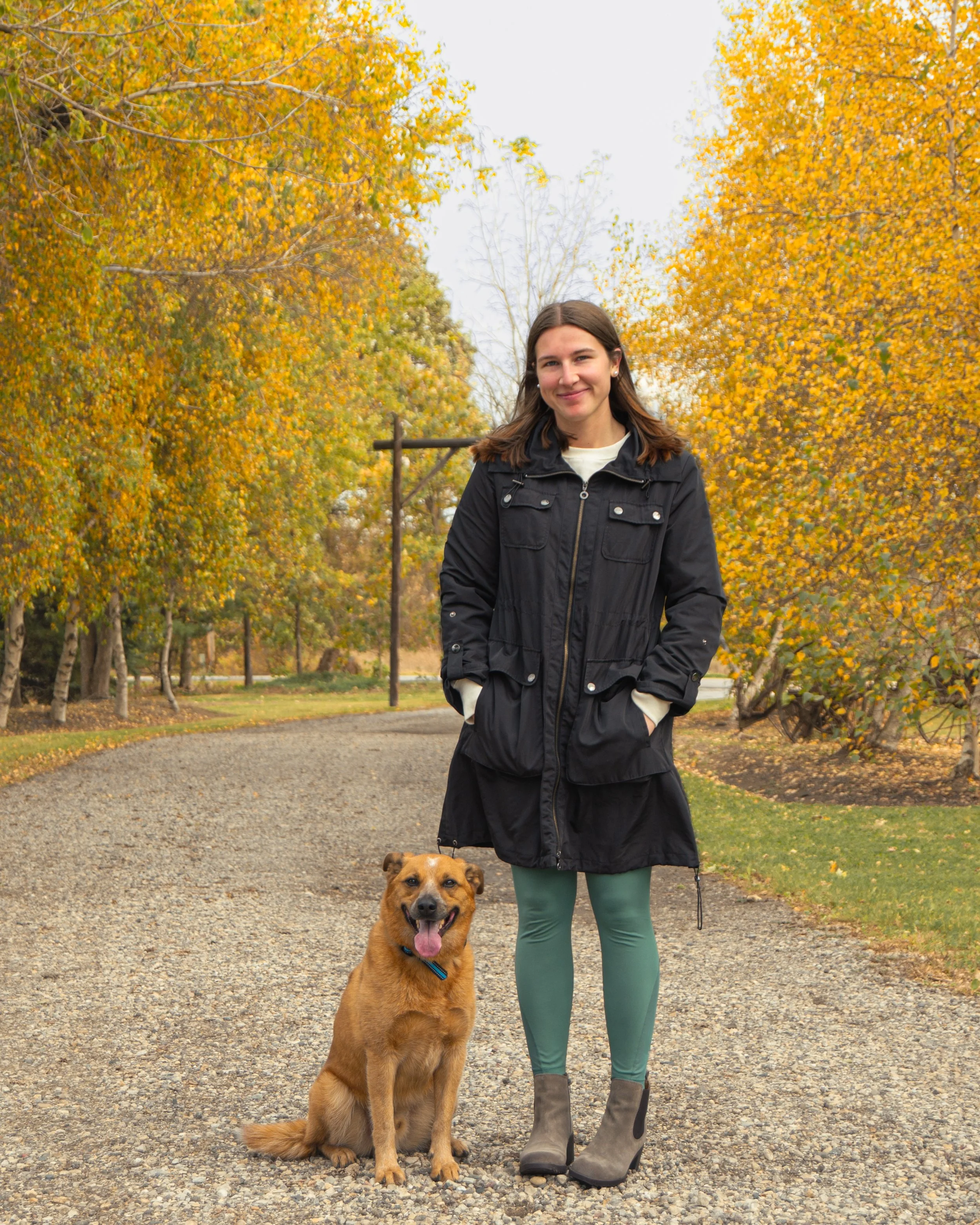 A young woman with long brown hair wearing a black coat, teal leggings, and gray ankle boots stands on a gravel path with her happy brown dog sitting beside her. The background features trees with autumn-colored leaves.