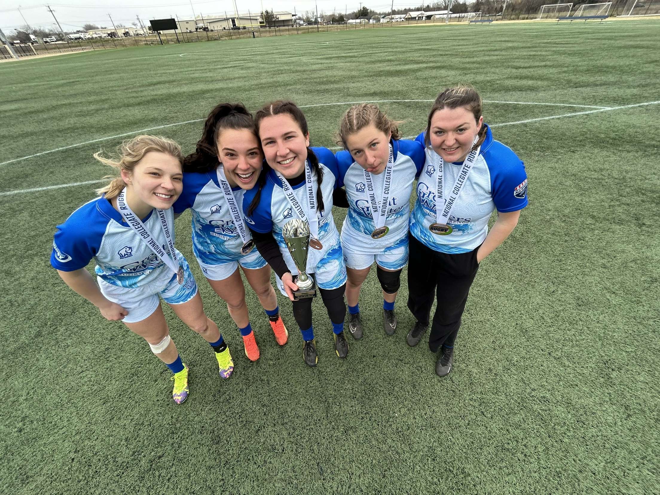 A group of five young women rugby players in blue and white uniforms, standing on a rugby field, smiling, with medals around their necks and a trophy at the center, celebrating a victory.