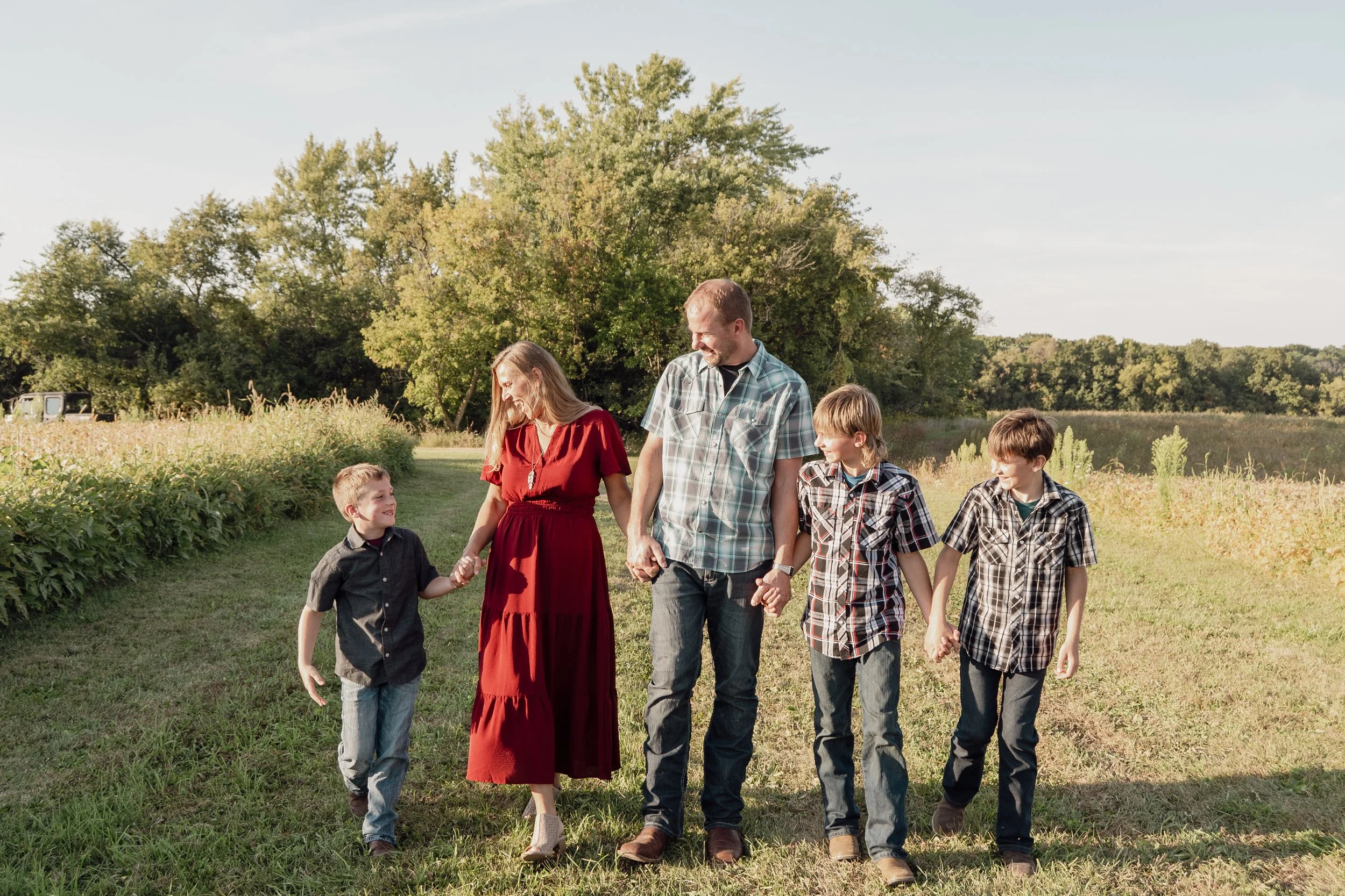 A family of five holding hands and walking along a grassy path in a rural field with trees in the background on a sunny day.