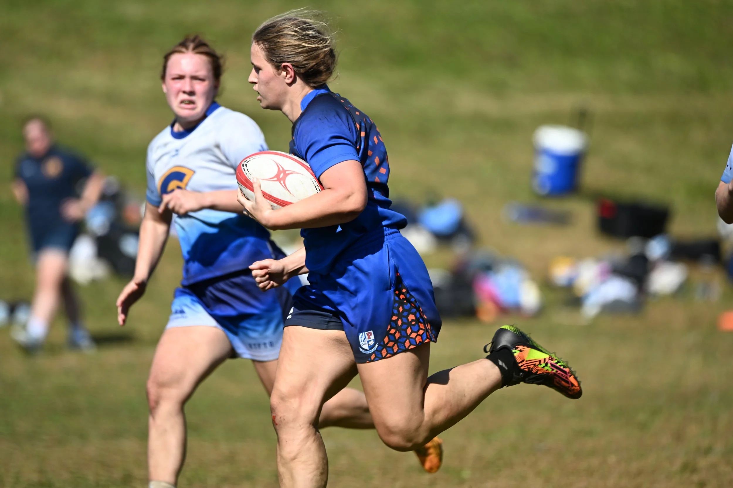 Female rugby players in blue uniforms playing on a grassy field, with one holding a white rugby ball with red details, and others running nearby, during a match.