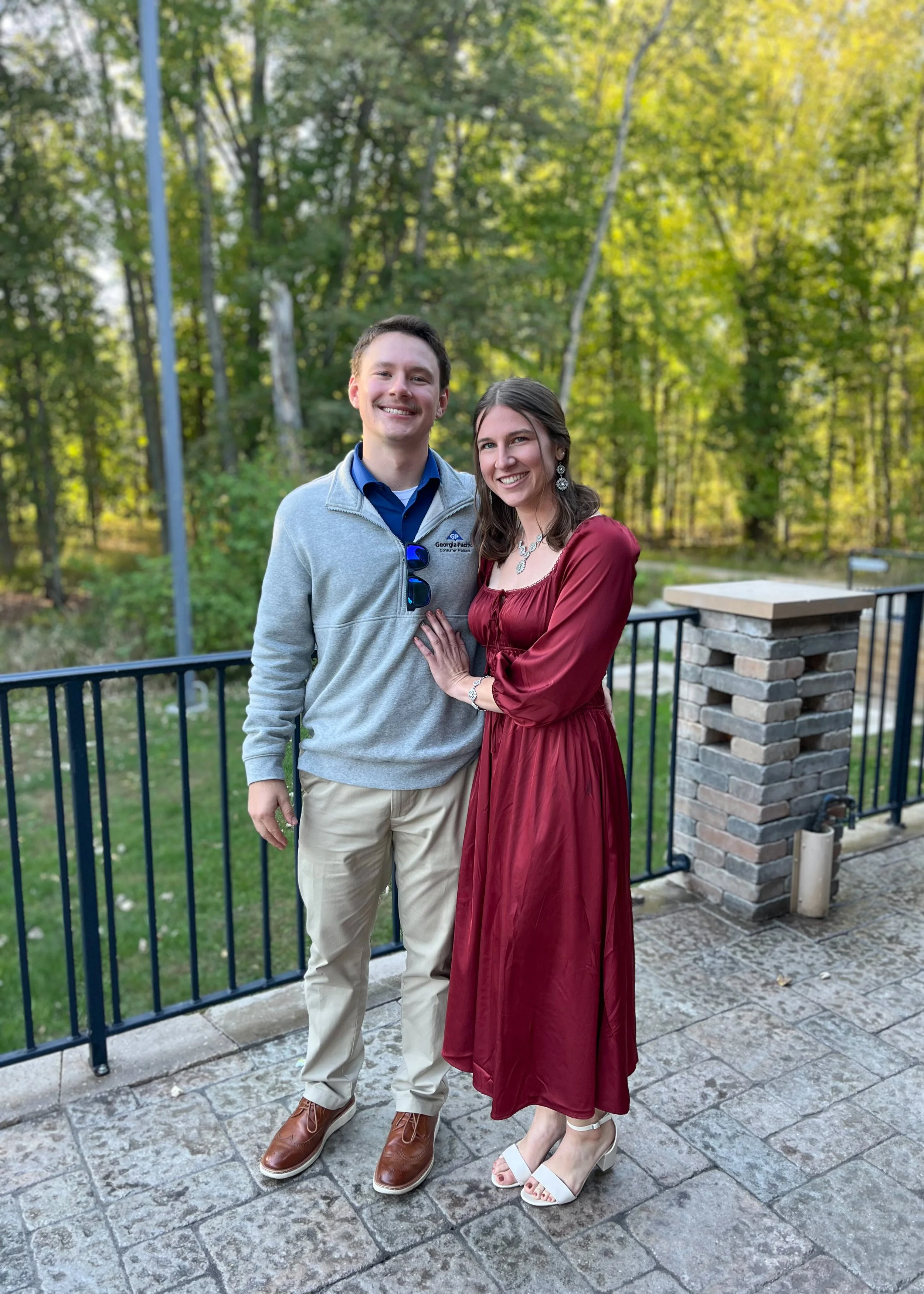 A young man and woman standing outside on a patio, smiling, with a wooded background, posing for a photo.