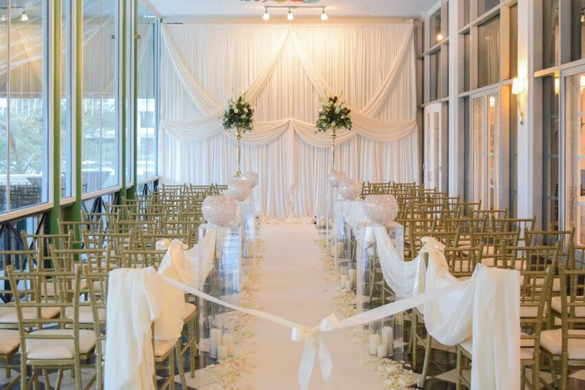 Elegant wedding ceremony setup inside a glass-enclosed venue with rows of chairs, floral arrangements, and draped white fabric creating a backdrop.