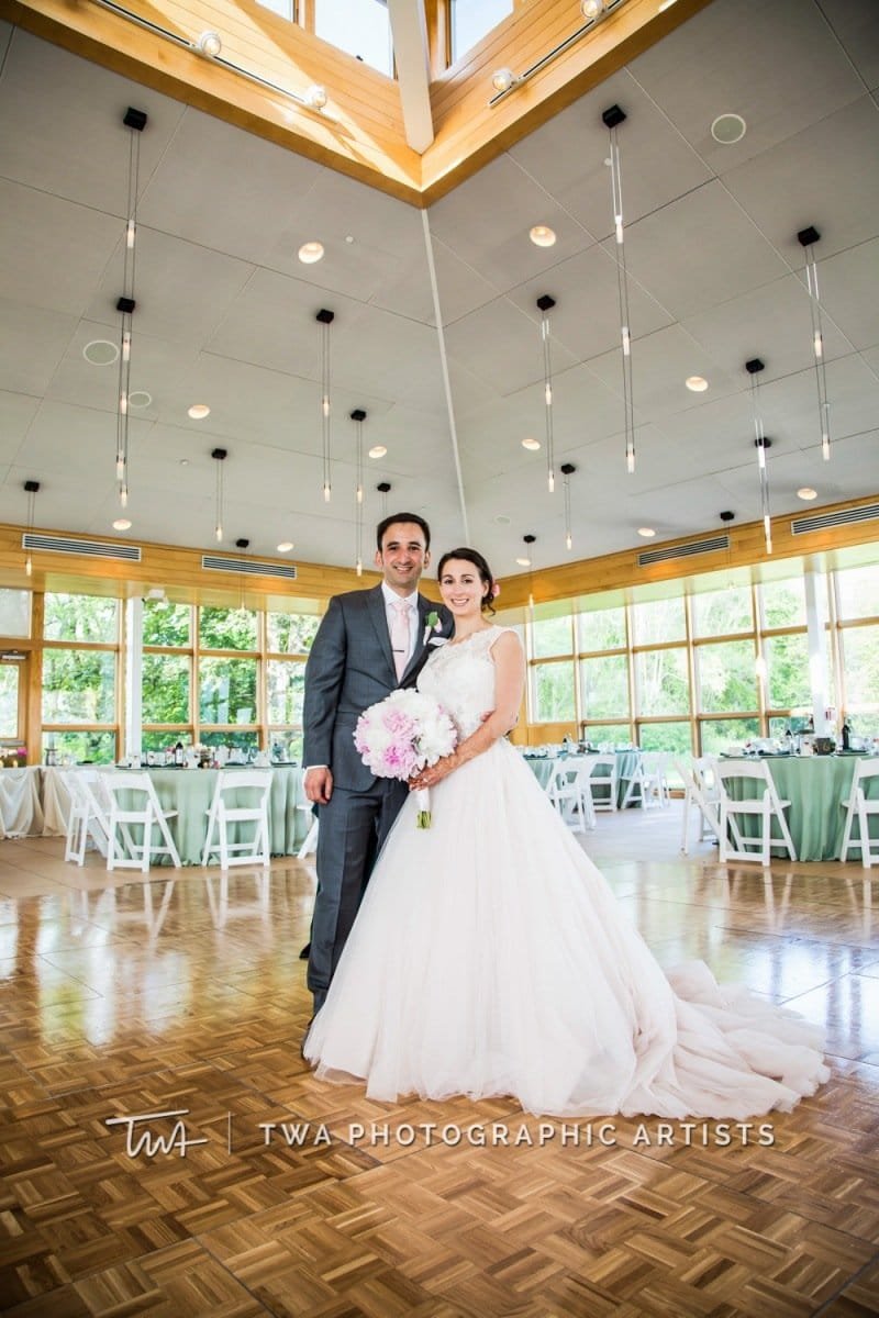 A bride and groom standing together indoors at a wedding reception, with the bride holding a bouquet of pink and white flowers, and the bride wearing a white wedding gown and the groom in a gray suit. The space has large windows, wooden accents, and modern hanging lights.