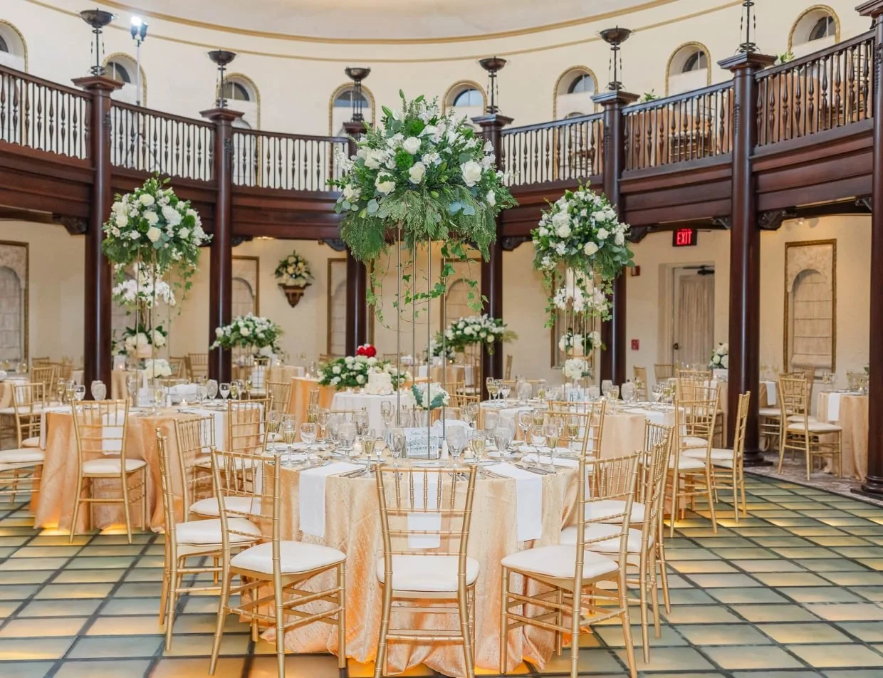 Elegant event venue with round tables draped in beige tablecloths, each set with glassware, silverware, and white napkins. Tall floral centerpieces with white flowers and greenery decorate each table. The venue features a second level with a wooden railing, wall-mounted flower arrangements, and a tiled floor.
