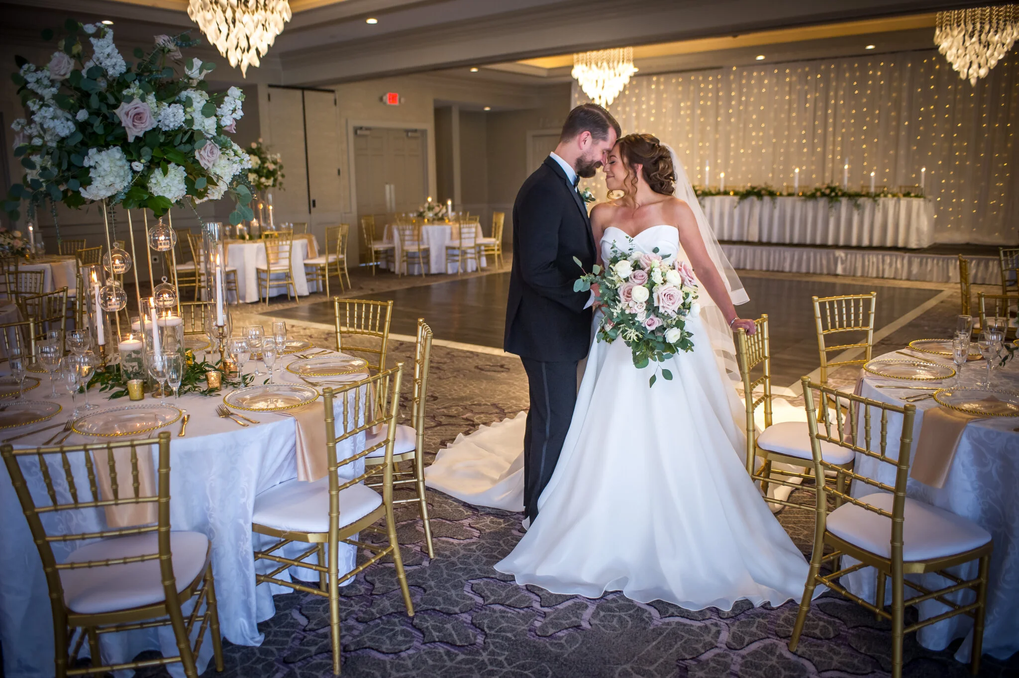 A bride and groom standing close together in a decorated wedding reception hall, with tables set for dinner and a large floral centerpiece nearby.