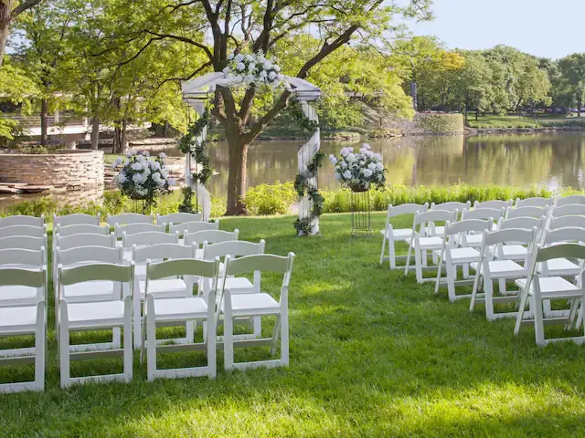 Outdoor wedding setup with white chairs facing a decorated arch by a river, surrounded by trees and grass.