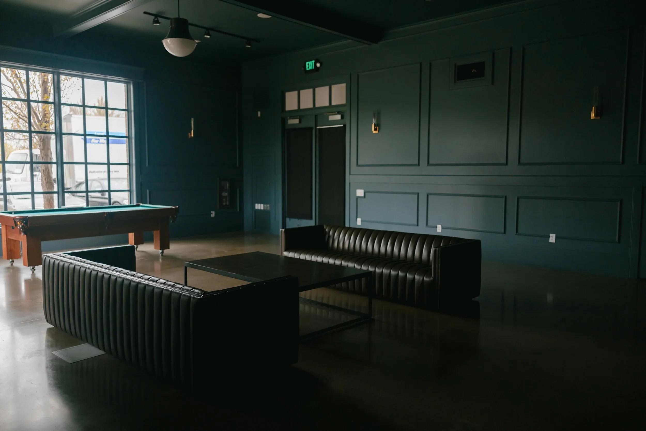 Interior of a dark-colored room with two black leather sofas, a pool table, large window letting in natural light, wall sconces, an exit sign, and dark wooden wall paneling.