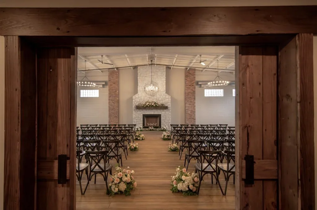 Inside a rustic wedding chapel with a fireplace, floral arrangements lining the aisle, and rows of black chairs facing the altar.
