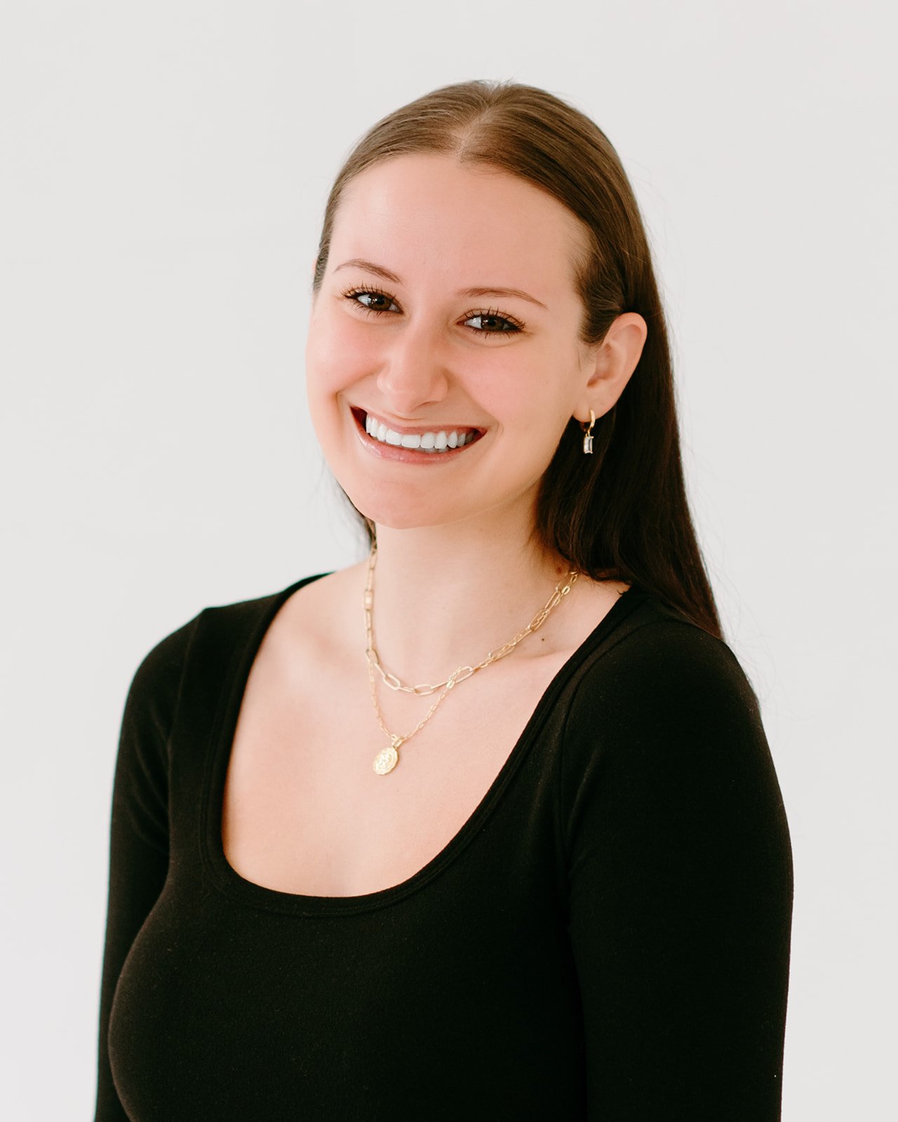 A young woman smiling, wearing a black top, gold jewelry, and earrings, with straight brown hair, against a plain light background.