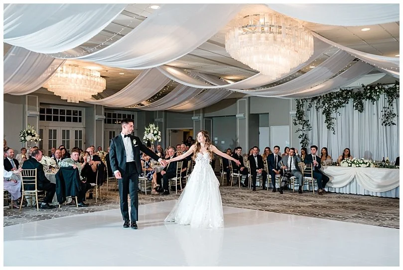 Bride and groom dancing at their wedding reception in a decorated ballroom with chandeliers and guests seated around the dance floor.