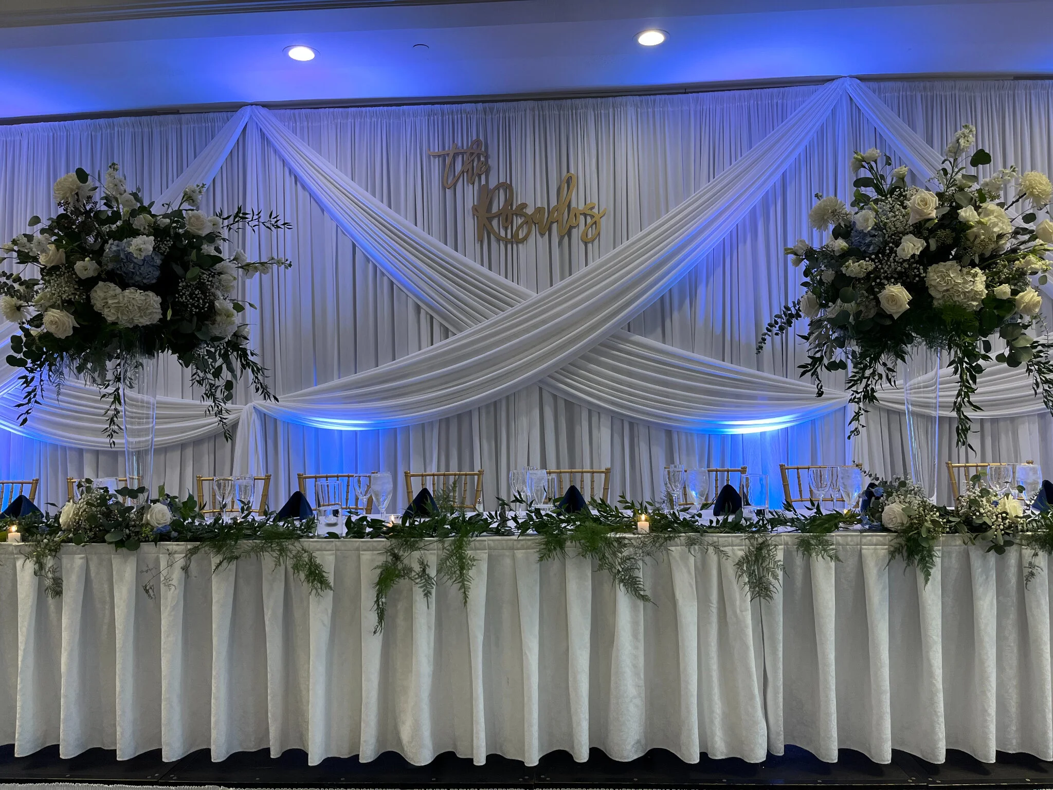 Decorated wedding reception table with white drapes, large flower arrangements, gold chairs, and a sign that says "the Rosados" on a white backdrop, with blue accent lighting.