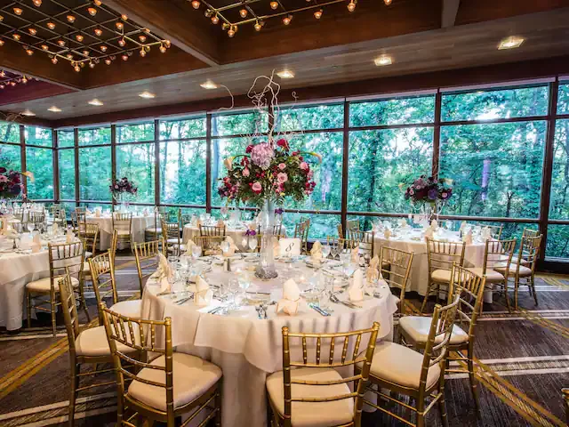 Round banquet table in a decorated event space with floral centerpieces and gold chairs, large windows with greenery outside.