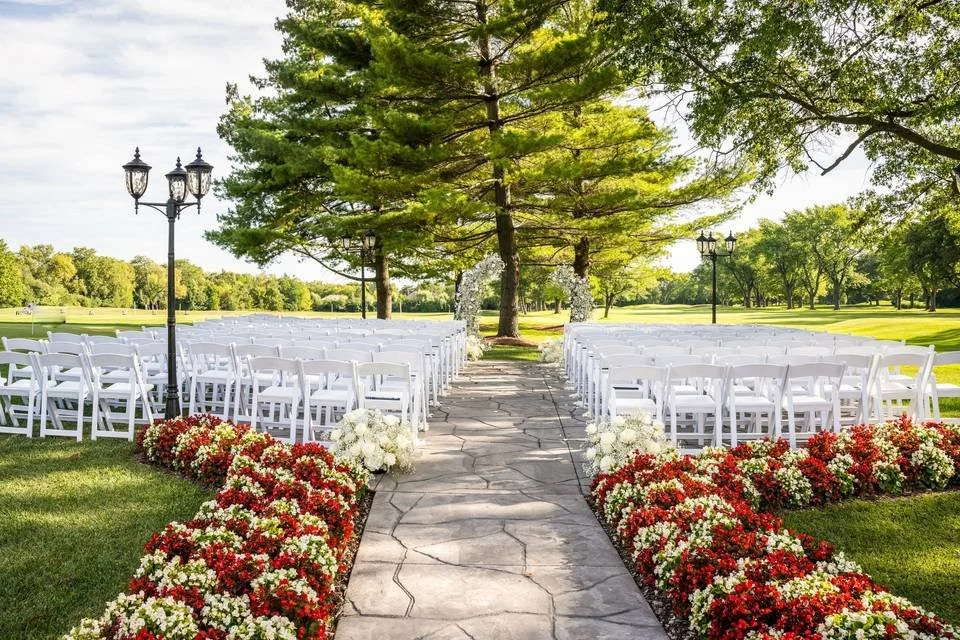 Outdoor wedding setup with white chairs on either side of a stone aisle, decorated with red and white flowers, on a grassy lawn with large trees and vintage street lamps.