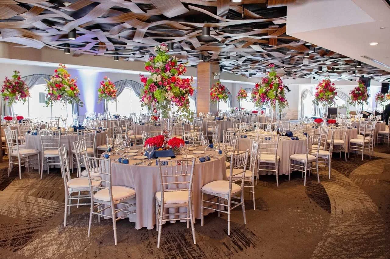 Elegant banquet hall decorated with large floral centerpieces, round tables with beige tablecloths, and white chairs, set up for a formal event.