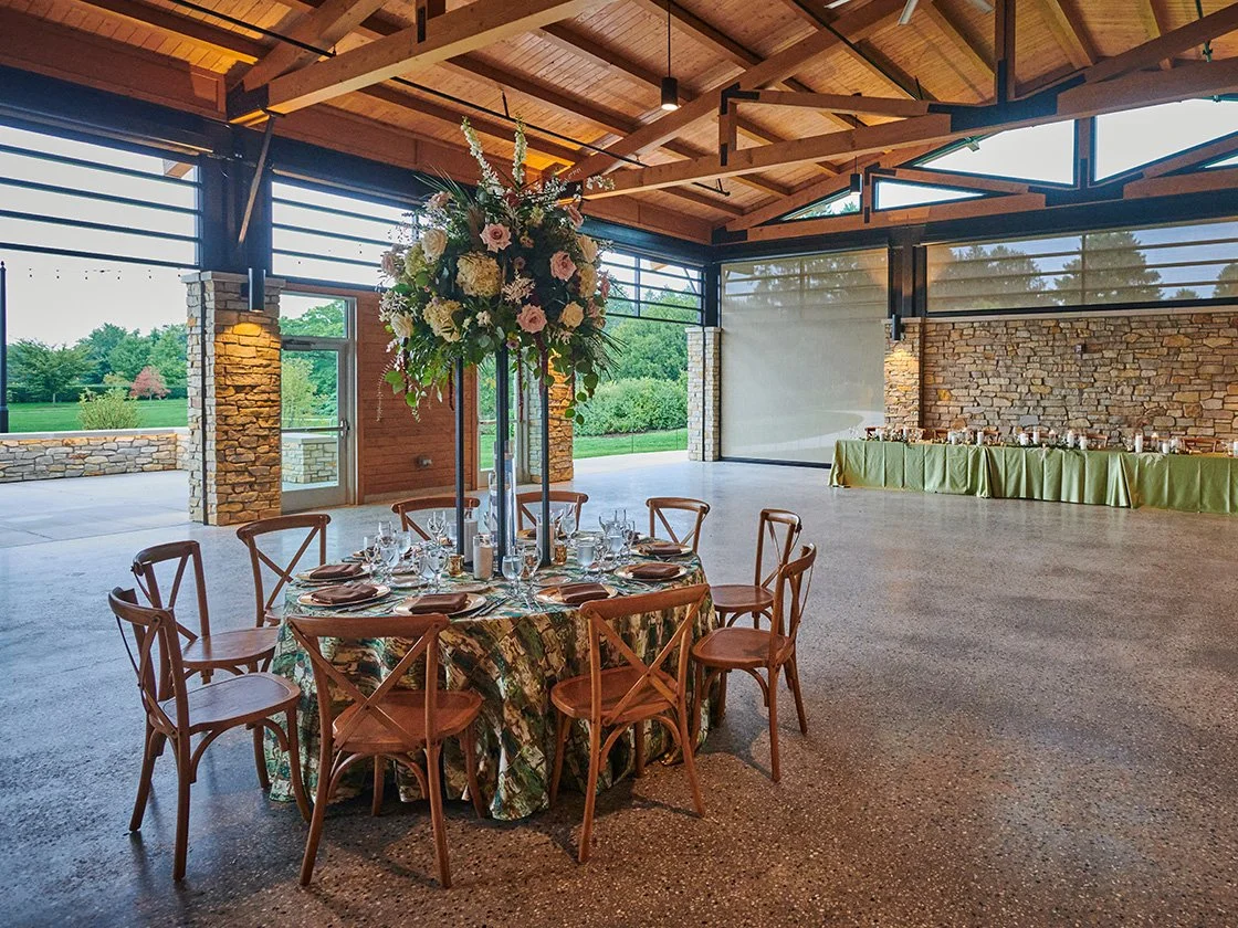 Indoor event space with a round table set for a meal, featuring a floral centerpiece, several glasses, and plates, with a long table along the wall in the background. Large windows overlook a green outdoor area.