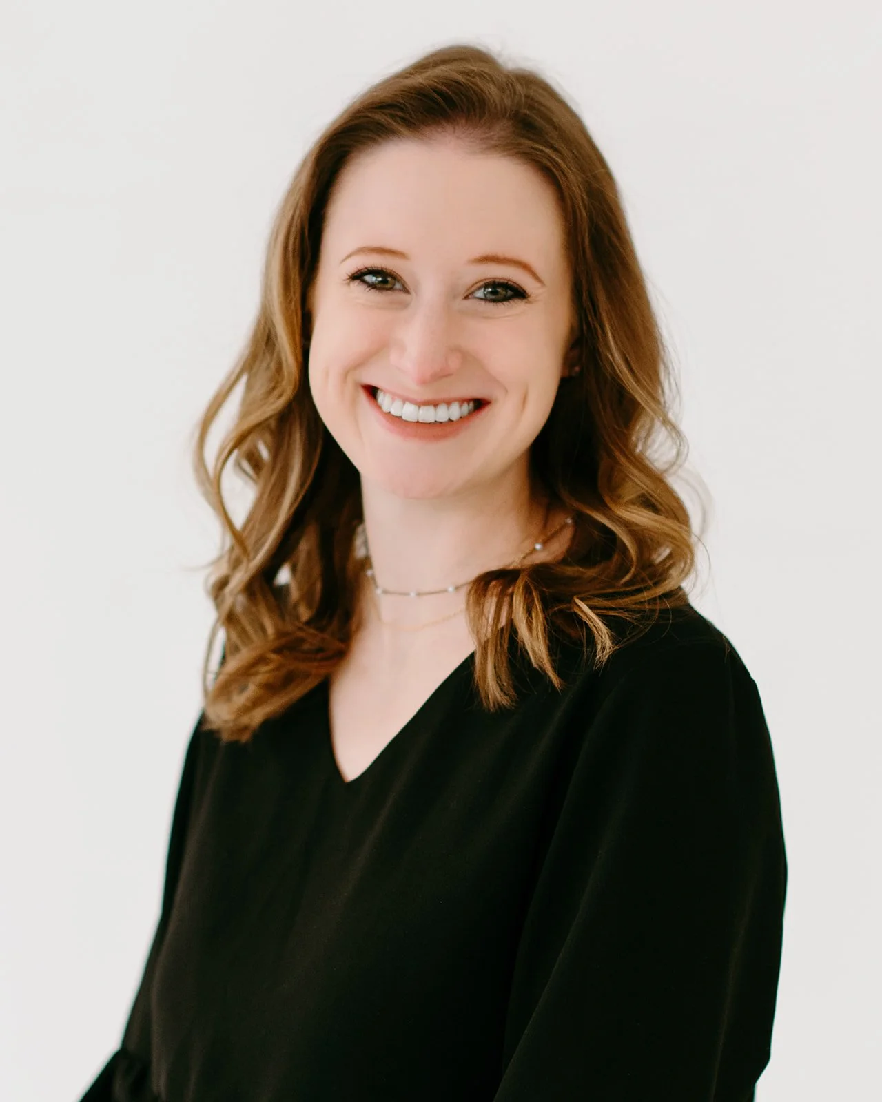 A woman with shoulder-length wavy auburn hair, smiling, wearing a black top and layered necklaces against a white background.