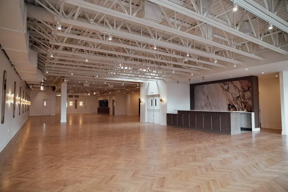 Spacious, empty event or gallery space with wooden flooring, white walls, and exposed ceiling beams, featuring wall-mounted lights and a marble accent wall.